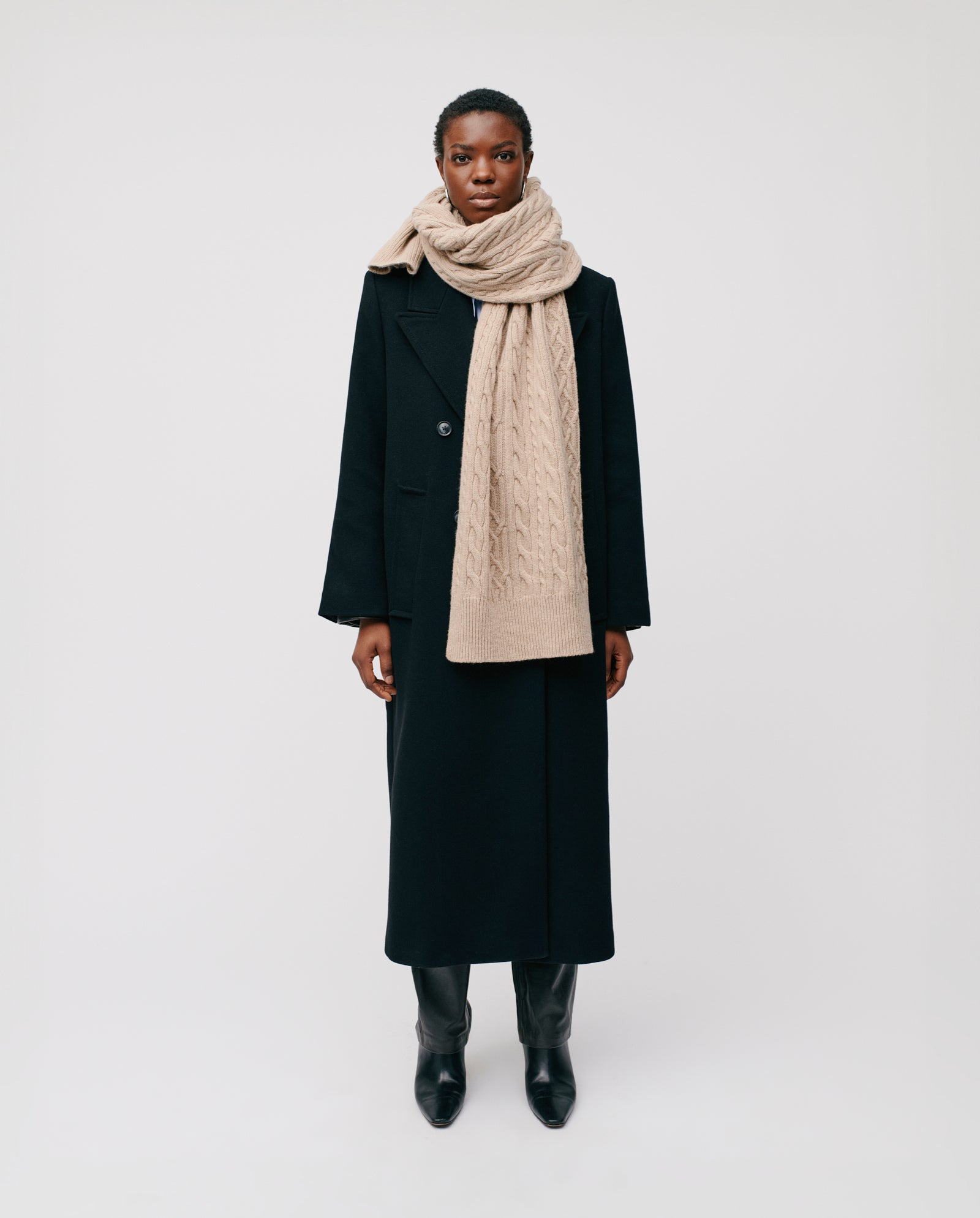 Person wears a long black coat, black boots, and the IVY OAK AMELIA Scarf—a chunky supergeelong extrafine merino wool cable knit—standing before a plain background. An ideal winter style choice.