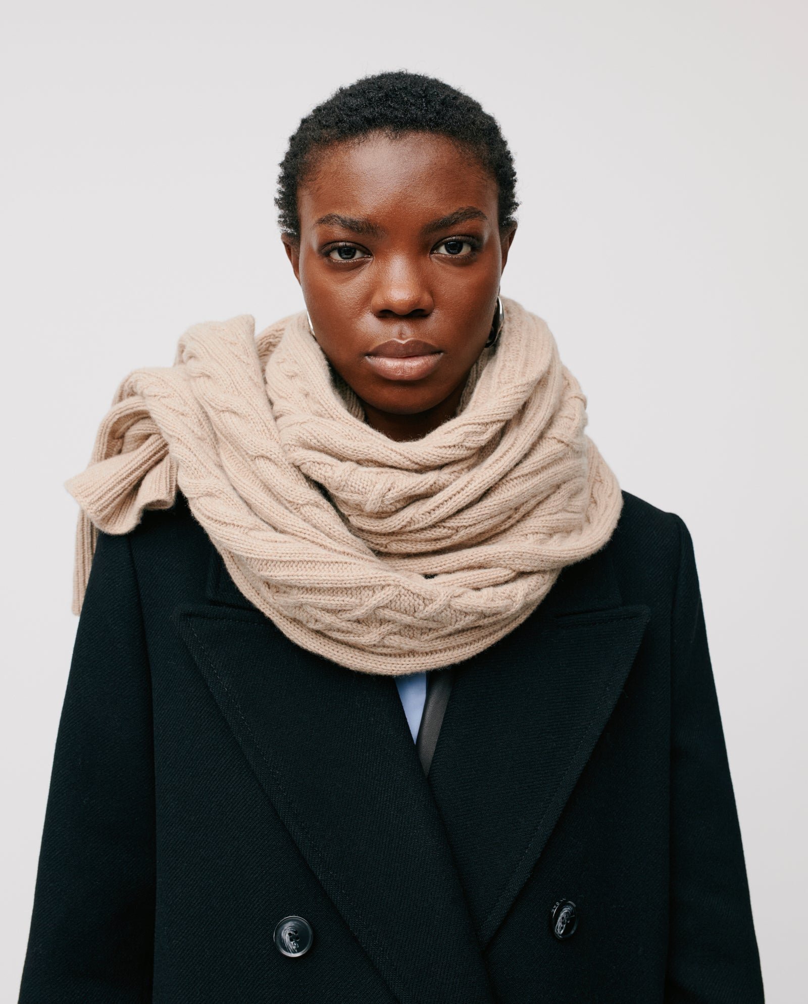 A person wears the IVY OAK AMELIA Scarf, a large beige cable knit crafted from supergeelong extrafine merino wool, styled with a black coat against a white background—ideal for chilly winter days.