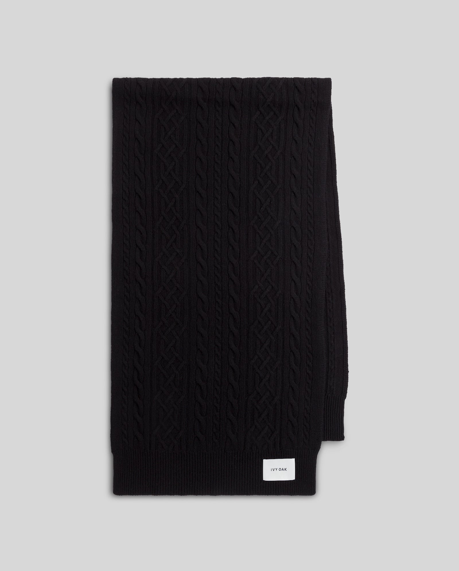 A folded black IVY OAK AMELIA Scarf in extrafine supergeelong merino wool cable knit, featuring a small white label, displayed on a light gray background.