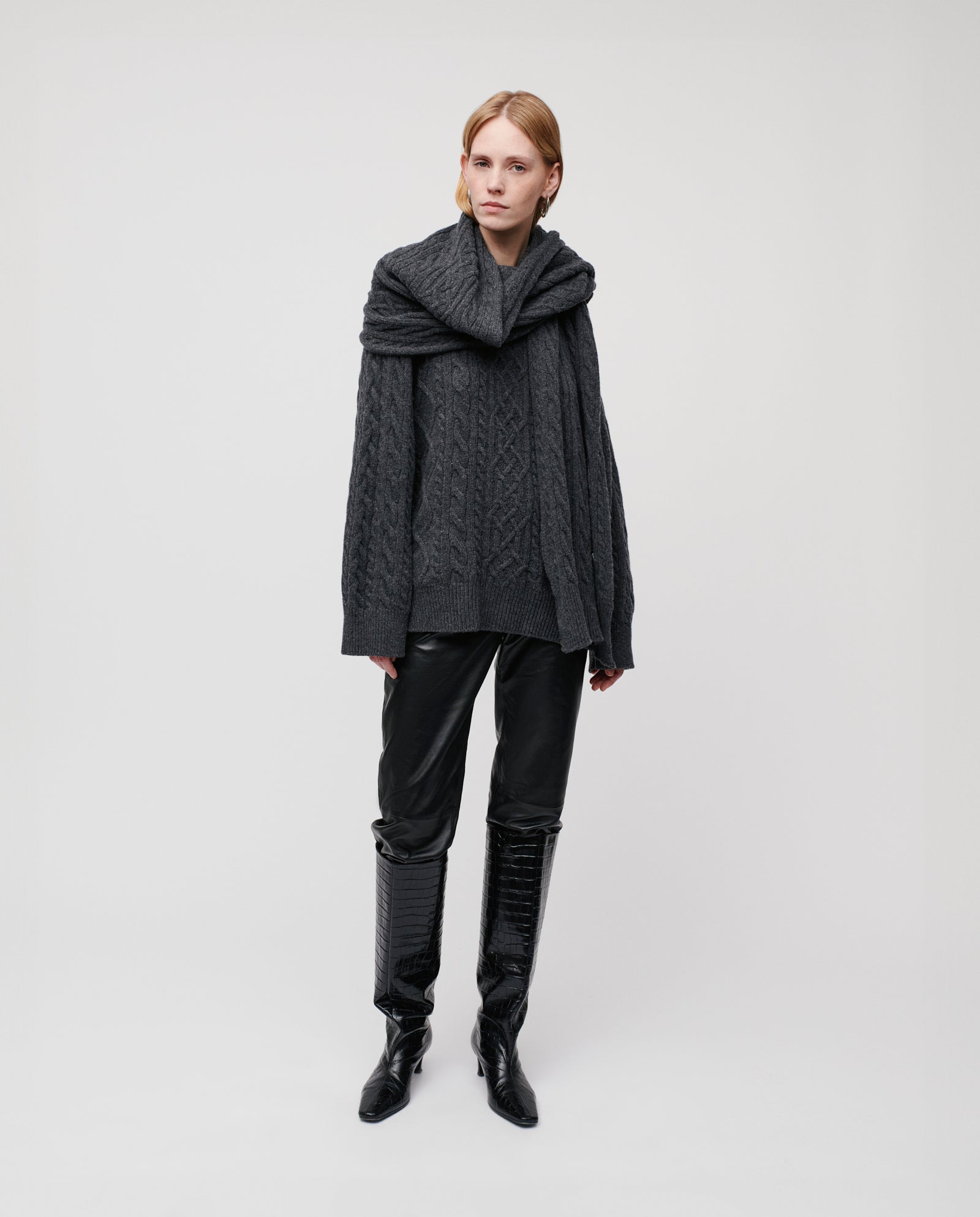 A person in a dark cable-knit sweater, black leather pants, knee-high black boots, and the IVY OAK AMELIA Scarf stands facing forward—ready for crisp winter days.