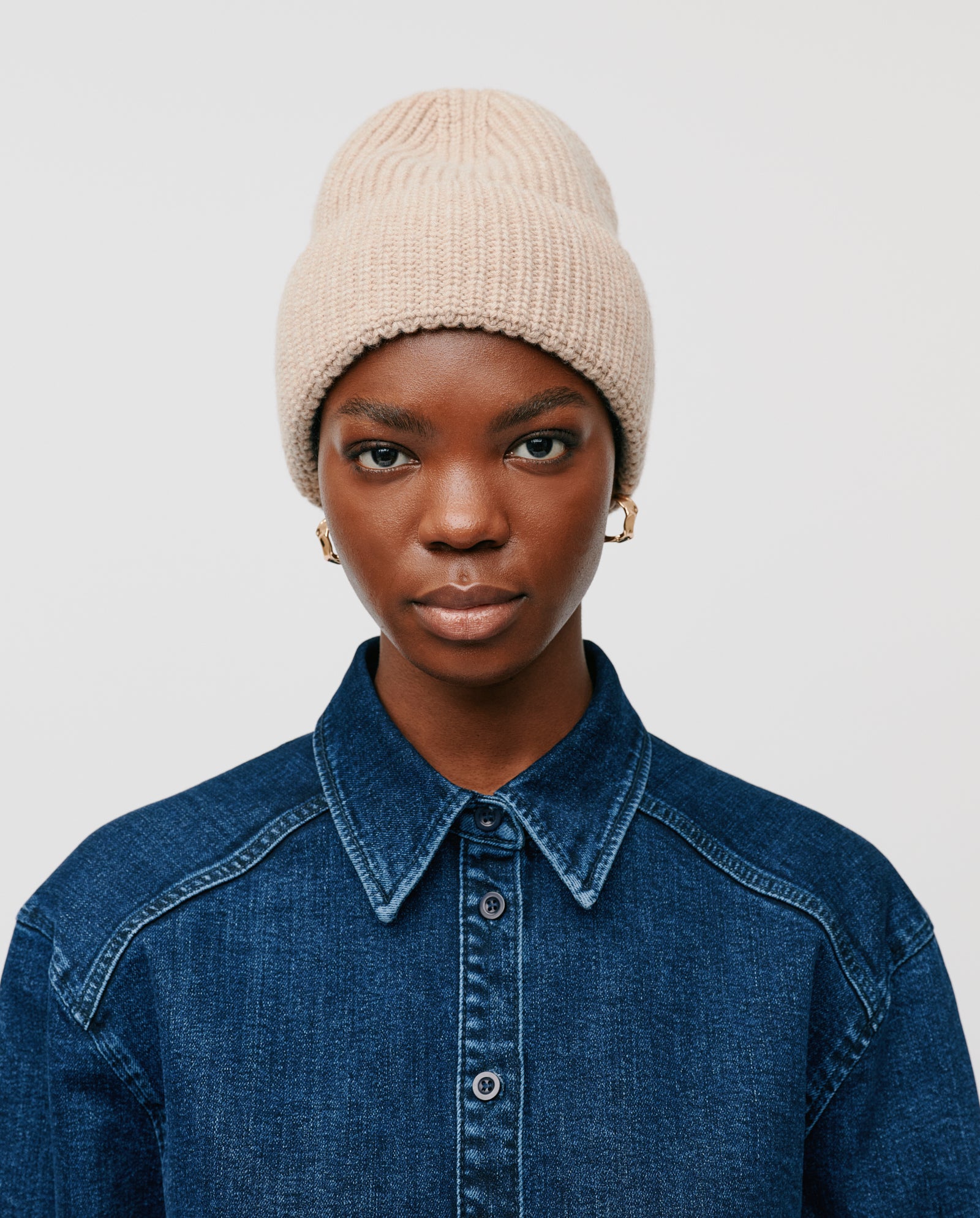 A person wears the IVY OAK AMBER Beanie in beige merino wool ribbed knit with a denim shirt, looking at the camera against a plain light background.
