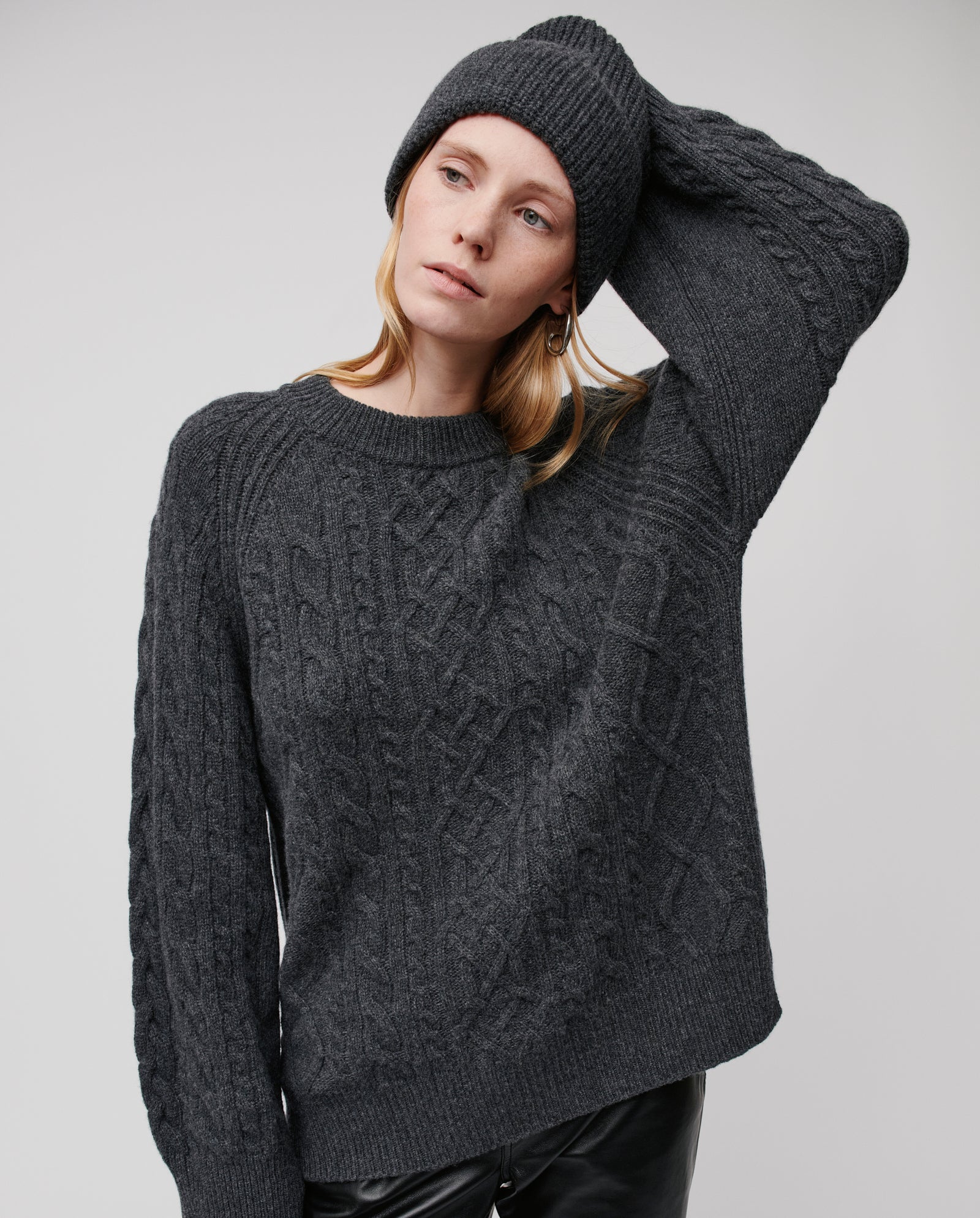A woman poses with one hand touching her hat, wearing the IVY OAK AMBER Beanie and a dark gray merino wool cable-knit sweater.