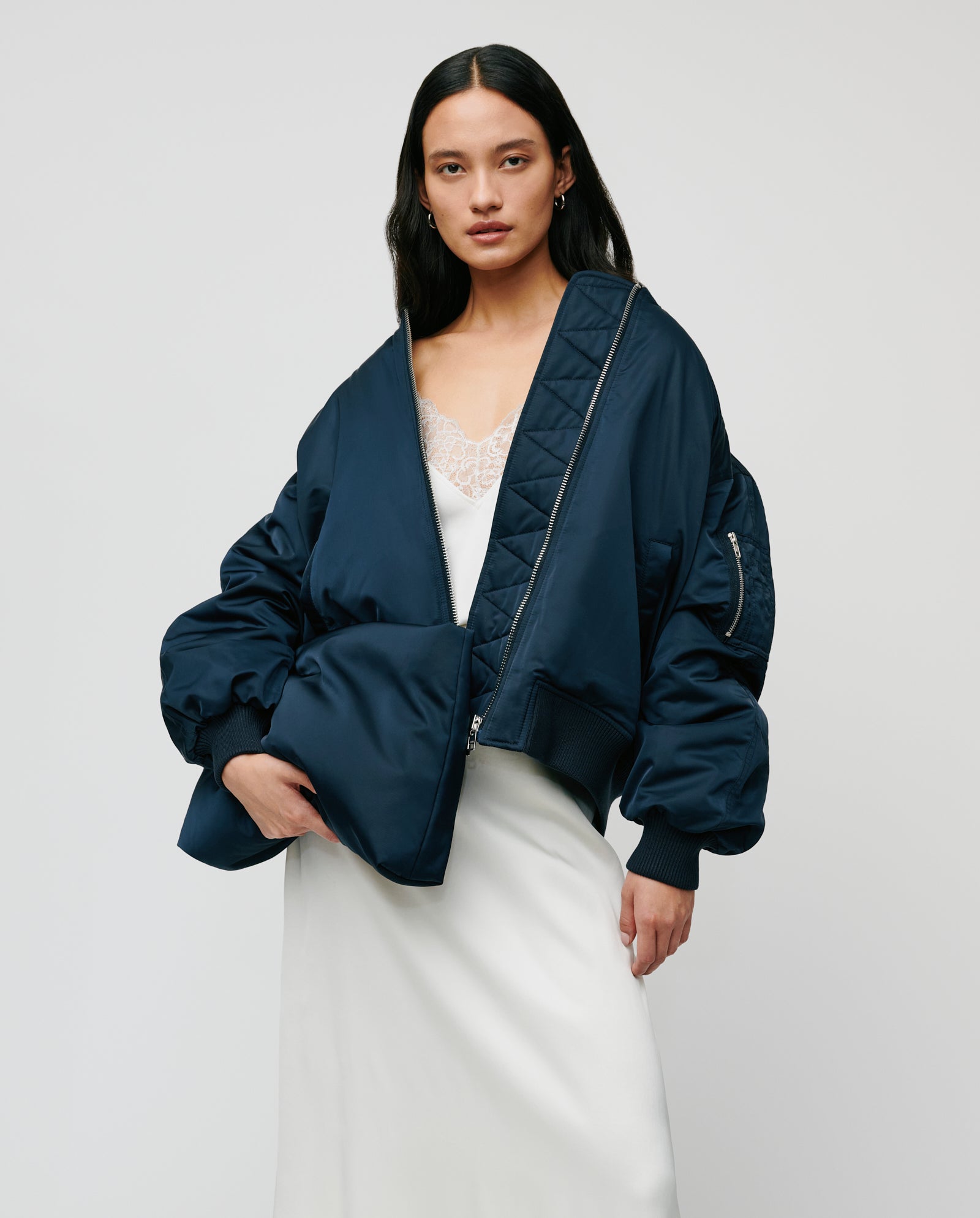 A woman stands against a plain background holding the IVY OAK AMINA ROSE Clutch, wearing a navy oversized bomber jacket with a metal zip over a white lace slip dress.
