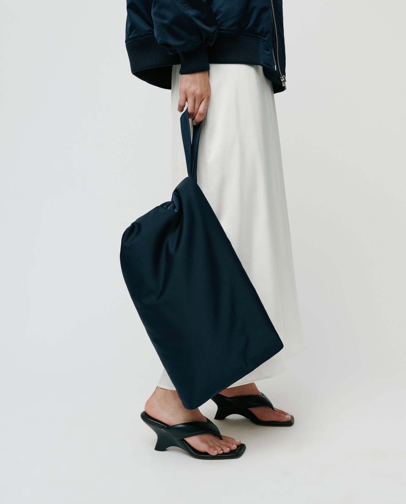 A person in a white skirt holds the IVY OAK AMINA ROSE Clutch, featuring a large padded design with a metal zip, paired with black heeled sandals against a plain background.