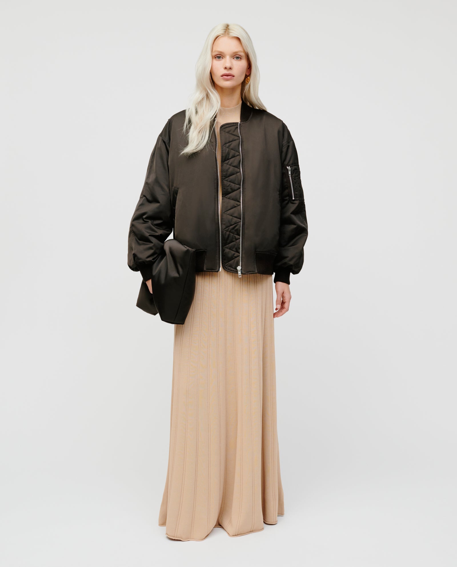 Woman in a long beige dress and oversized black bomber jacket holds the IVY OAK AMINA ROSE Clutch, featuring padding and a metal zip, against a plain white background.