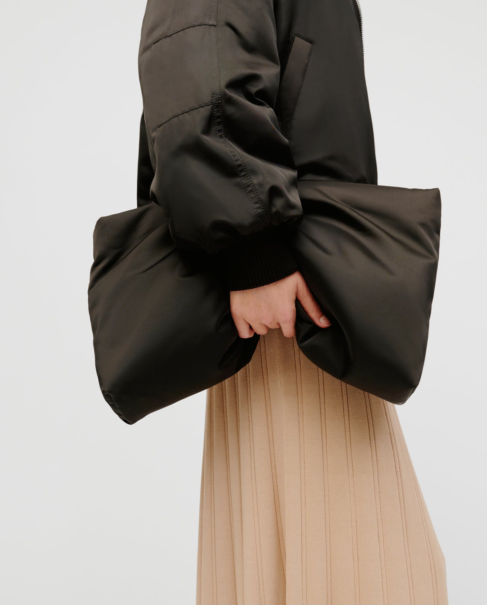 A person holds the hem of a beige ribbed skirt and showcases the IVY OAK AMINA ROSE Clutch, styled with a shiny black puffer jacket, against a plain background.