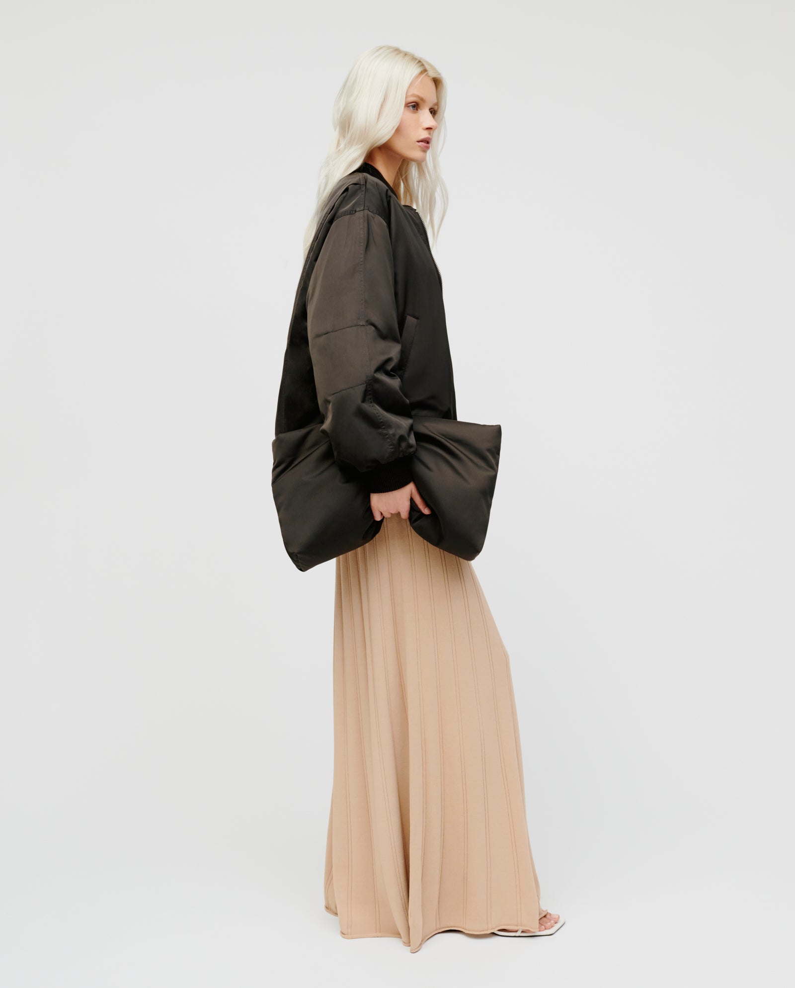 Woman with long blonde hair stands sideways in a black oversized jacket and beige pleated maxi skirt, holding the IVY OAK AMINA ROSE Clutch, a padded bag with a metal zip.