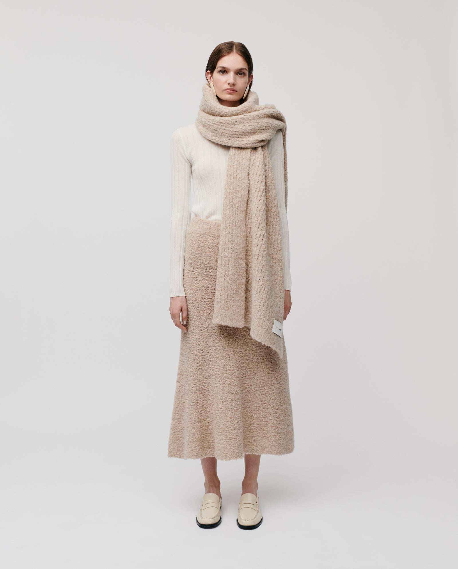 A woman in a cream sweater, beige bouclé skirt, and cream loafers stands against a white background, styled with the large ALINA Scarf by IVY OAK—an ideal cozy winter look.