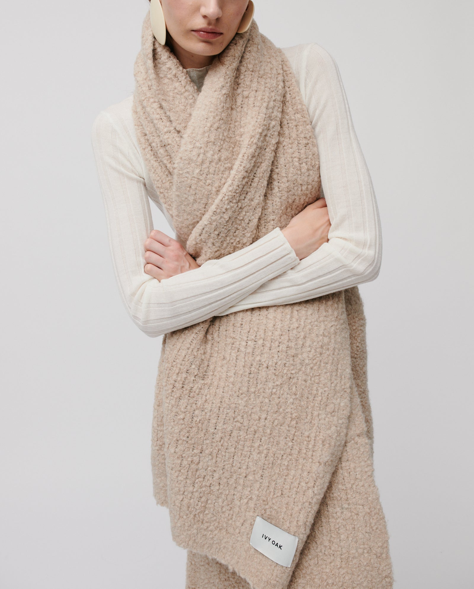 A woman in a white ribbed top wears the ALINA Scarf by IVY OAK, crafted from soft bouclé yarn with a visible brand label—perfect for cozy winter days.