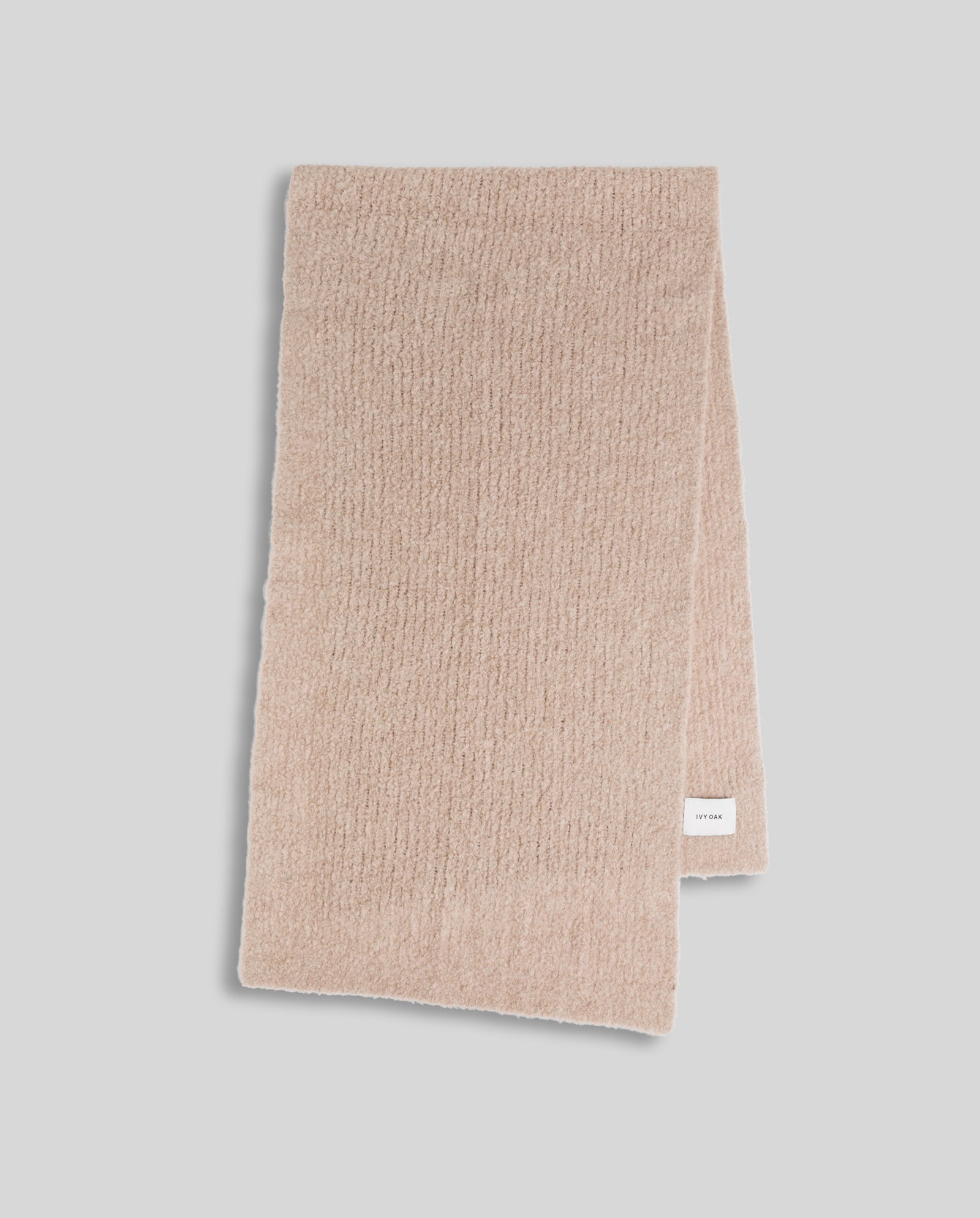 The ALINA Scarf by IVY OAK is shown folded on a light gray background, featuring cozy beige bouclé yarn and a small white label on one end.