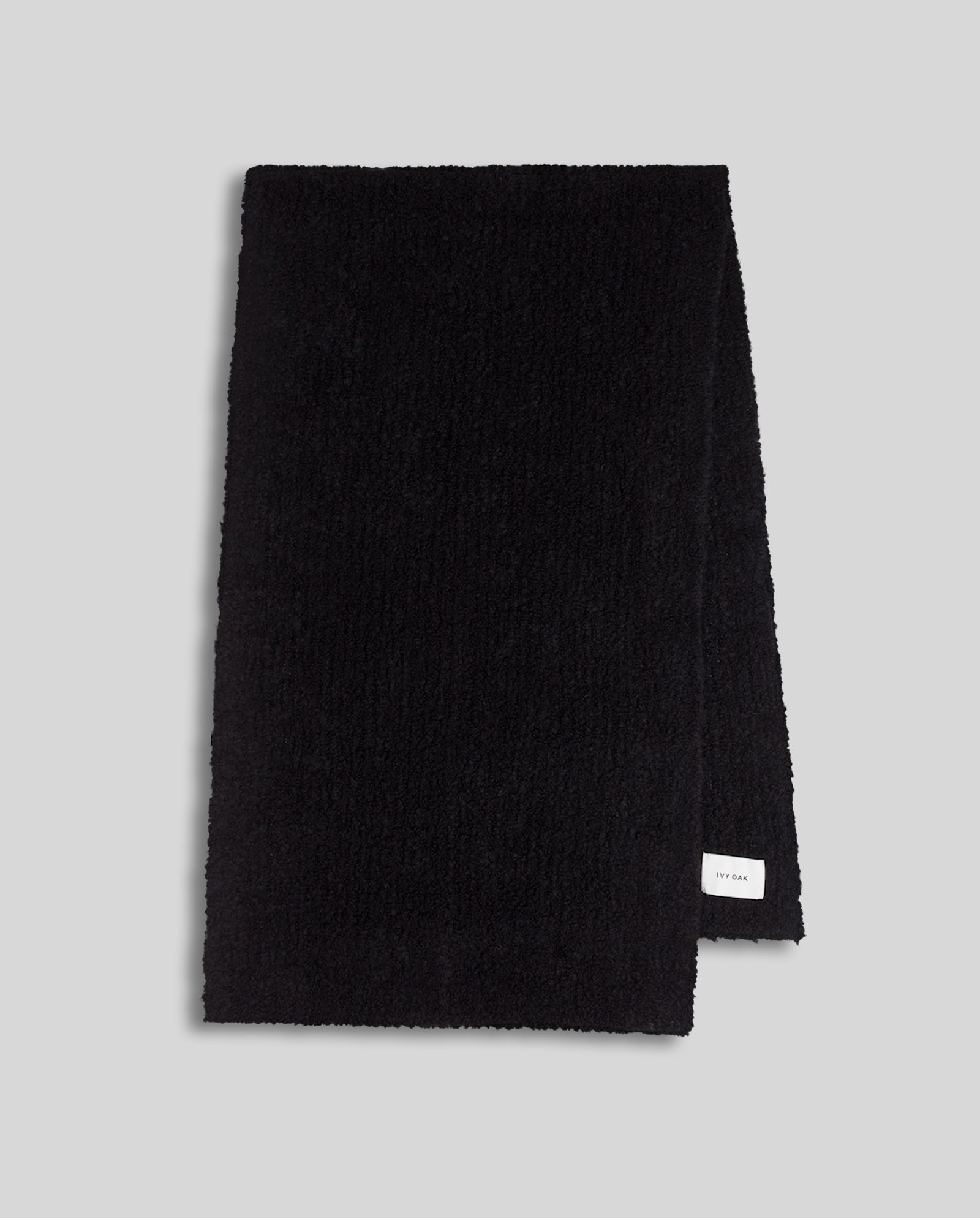 The ALINA Scarf by IVY OAK is a folded black winter scarf crafted from soft bouclé yarn, featuring a small white label on the bottom right, displayed on a light gray background.