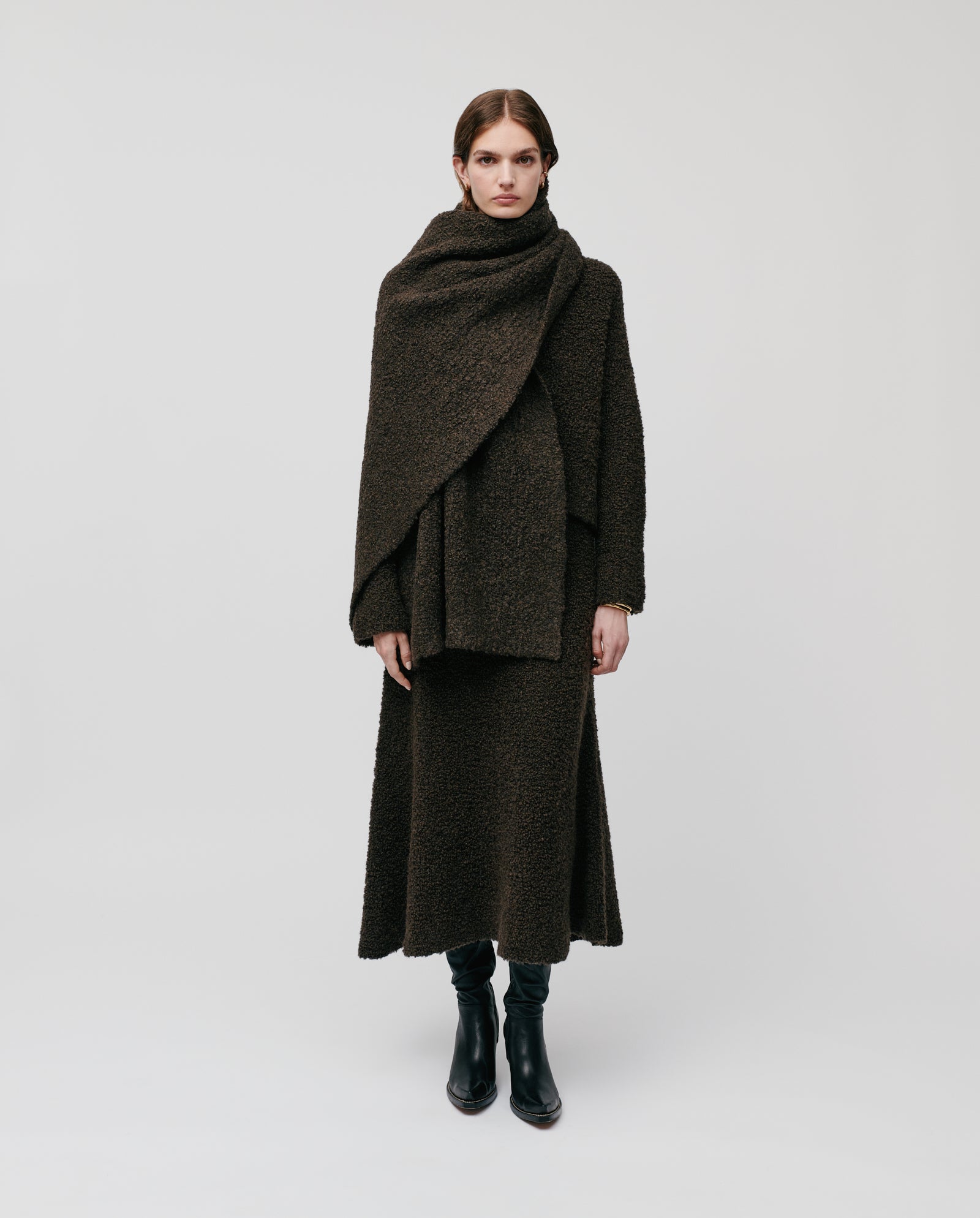 A person in a dark long coat with the ALINA Scarf by IVY OAK, both crafted from bouclé yarn, paired with black boots and set against a plain background—an ideal look for chilly winter days.