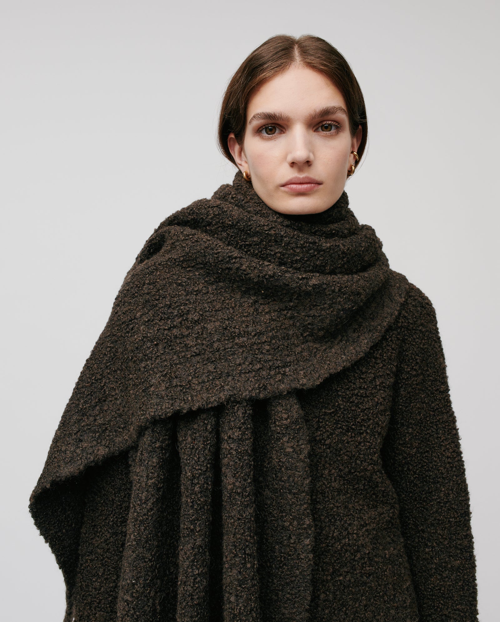 Woman wears the ALINA Scarf by IVY OAK—a large, textured dark brown bouclé yarn accessory—paired with a matching coat, looking at the camera against a plain background. Ideal for cozy winter days.