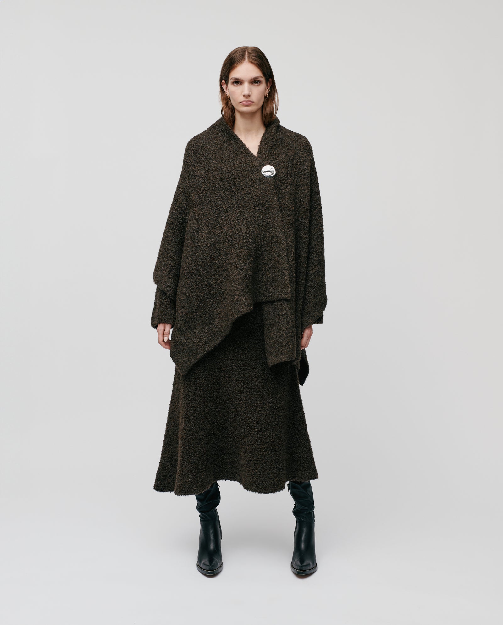 A woman stands against a light background in a dark textured cape and skirt with black boots, accessorized with the IVY OAK ALINA Scarf—long, wide, and crafted from bouclé yarn—ideal for chilly winter days.