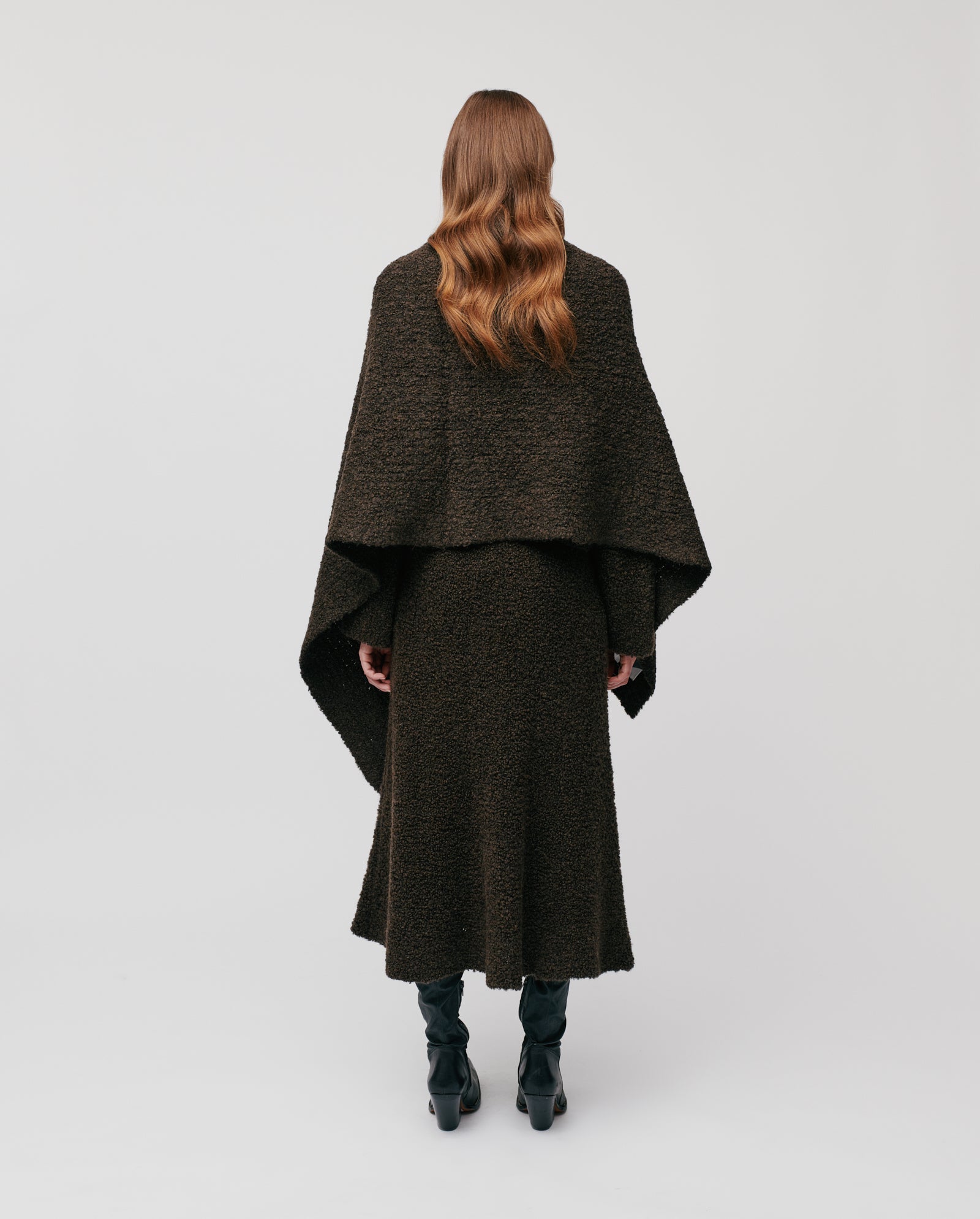 A woman with wavy hair wears a dark, textured cape and skirt, complemented by the long and wide ALINA Scarf from IVY OAK—perfect for winter days—as she stands with her back to the camera.