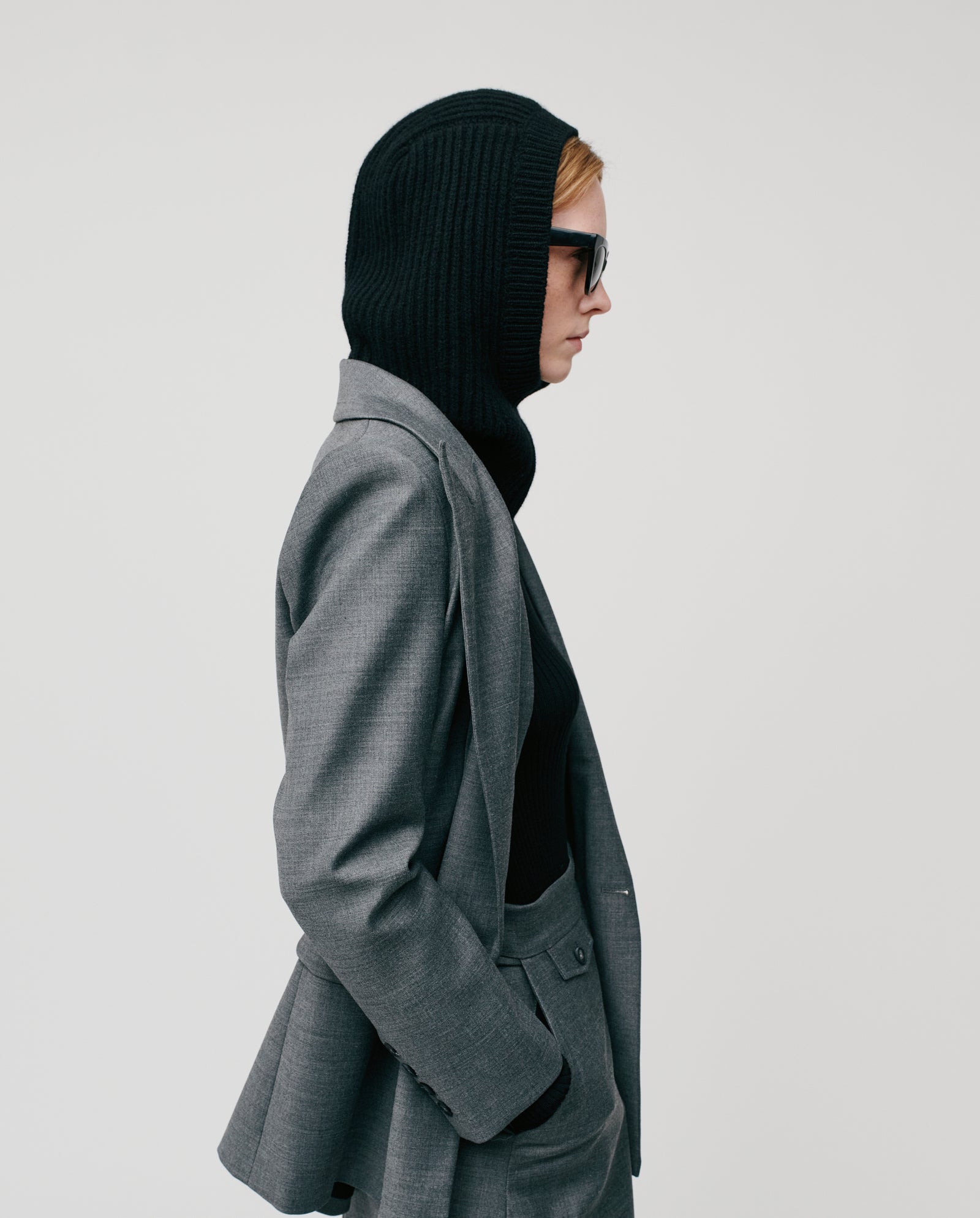 A person in profile wears a gray suit, IVY OAK ANNIE LEE Balaclava in extrafine merino wool rib, and sunglasses against a plain light background.
