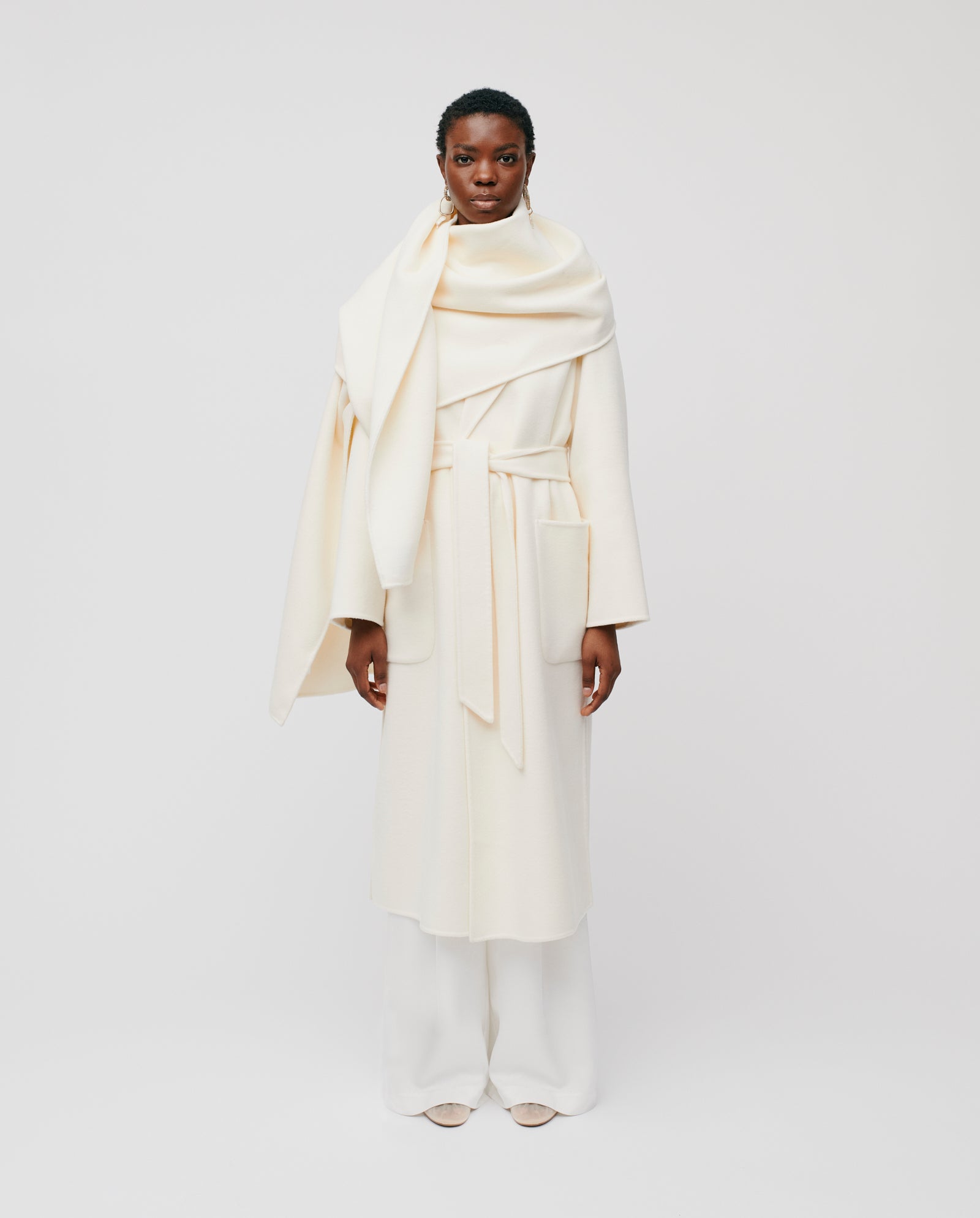 A person styles a cozy look with the IVY OAK AMY Wool Scarf, a long cream coat, white pants, and light shoes.