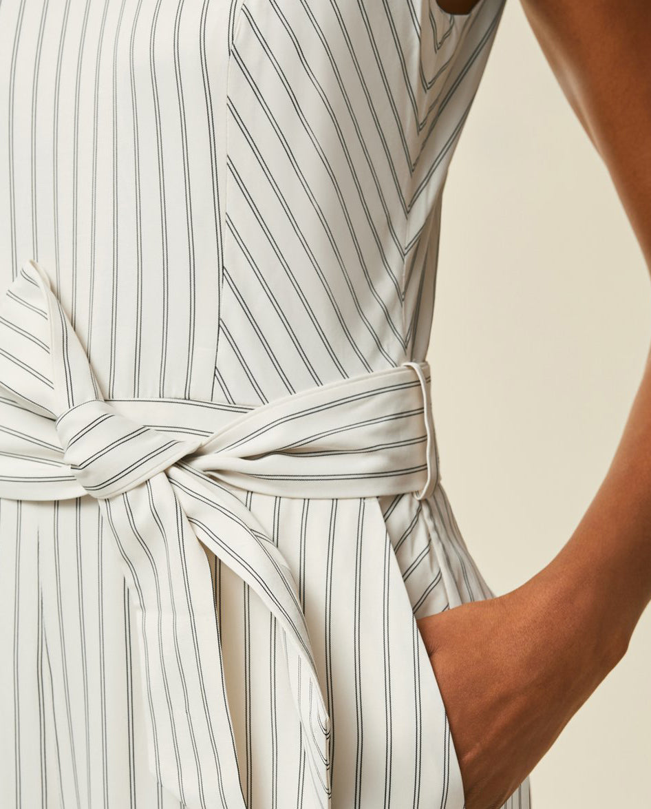 Close-up of a person wearing a white sleeveless dress with thin black stripes, a tied belt, and hand in pocket.