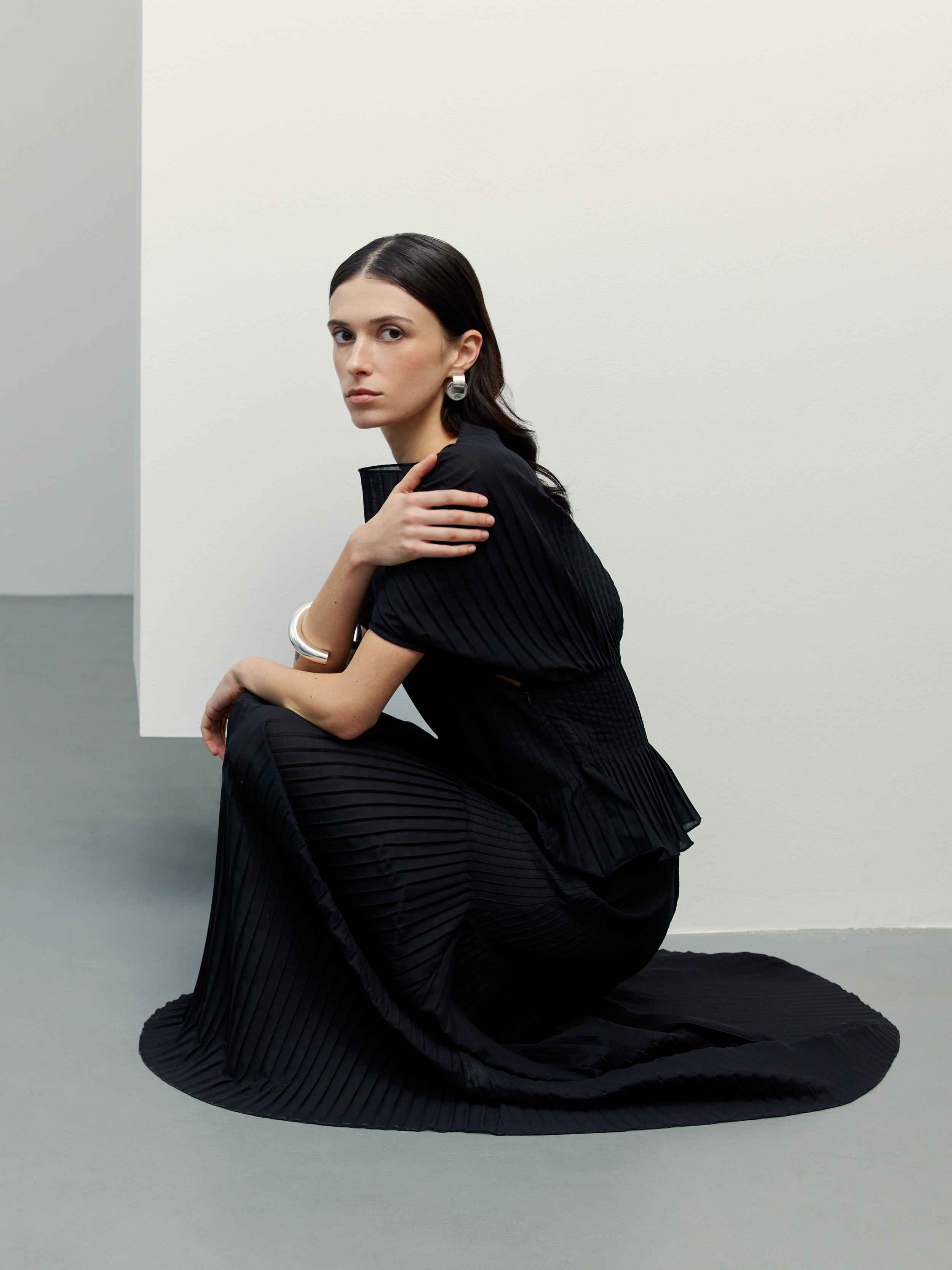 Woman in a pleated black dress sits on the floor against a minimalist white and gray background.