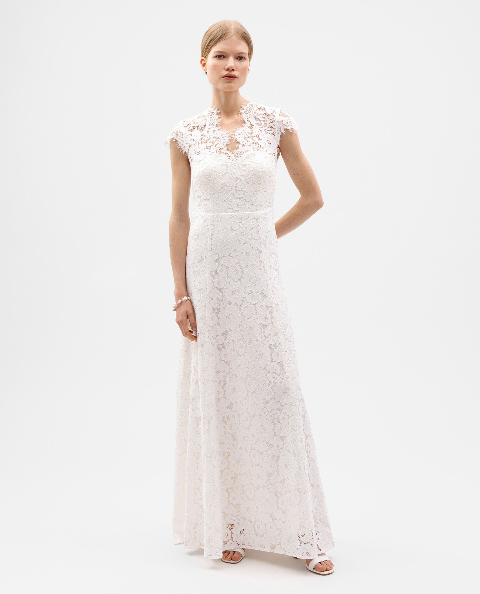 A woman wears the IVY OAK DANIELLA Bridal Dress—a floor-length, form-fitting white gown with cap sleeves and a flared skirt in exquisite cotton blend lace—standing against a plain white background.