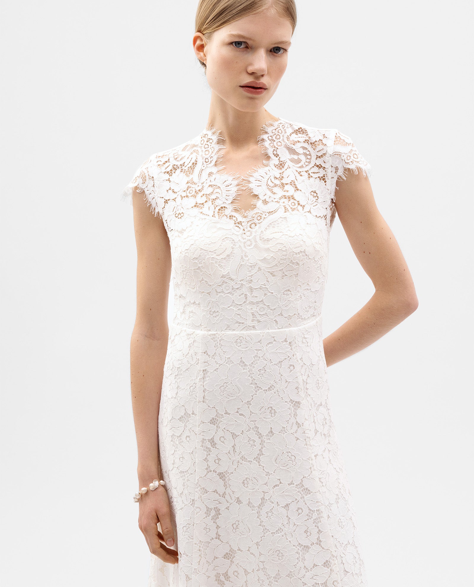A woman models the IVY OAK DANIELLA Bridal Dress, featuring a white cotton-blend lace design with cap sleeves, a high neckline, and a softly flared skirt, set against a plain background.