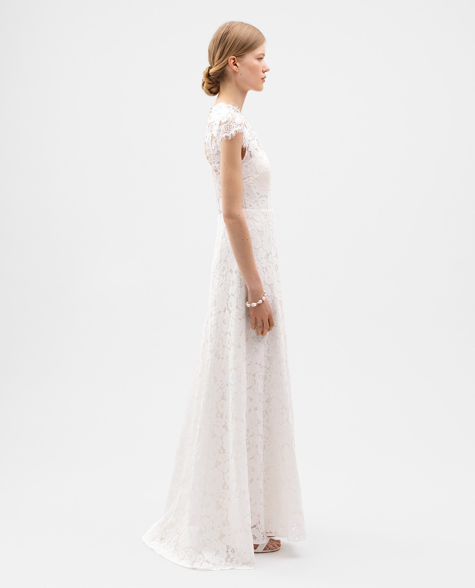 A woman in the IVY OAK DANIELLA Bridal Dress—a white cotton-blend lace, form-fitting short-sleeve gown—stands in profile against a plain white background.