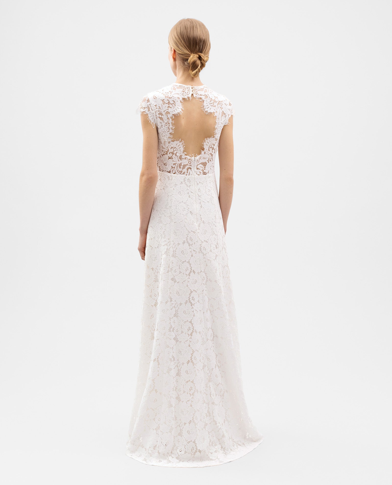 A woman stands facing away, wearing the IVY OAK DANIELLA Bridal Dress—a form-fitting white cotton-blend lace gown with an open back and flared skirt.