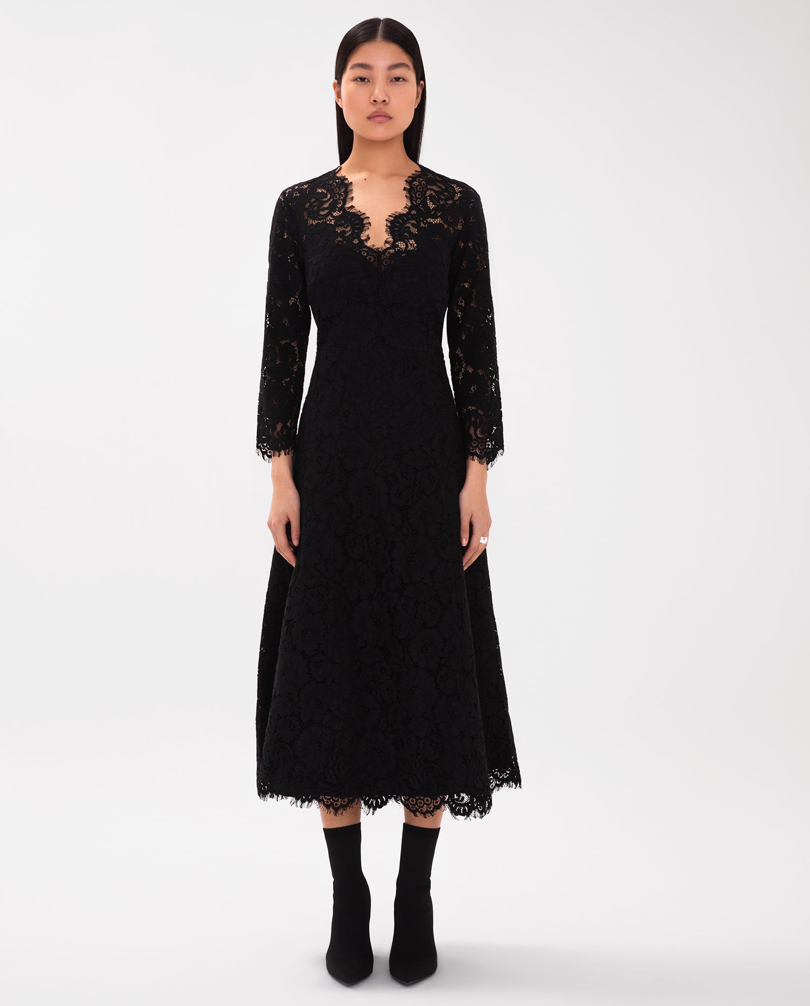 A woman wears the IVY OAK Flared Midi Lace Dress, featuring long sleeves, black lace, a V-neckline, flared skirt, and black ankle boots.