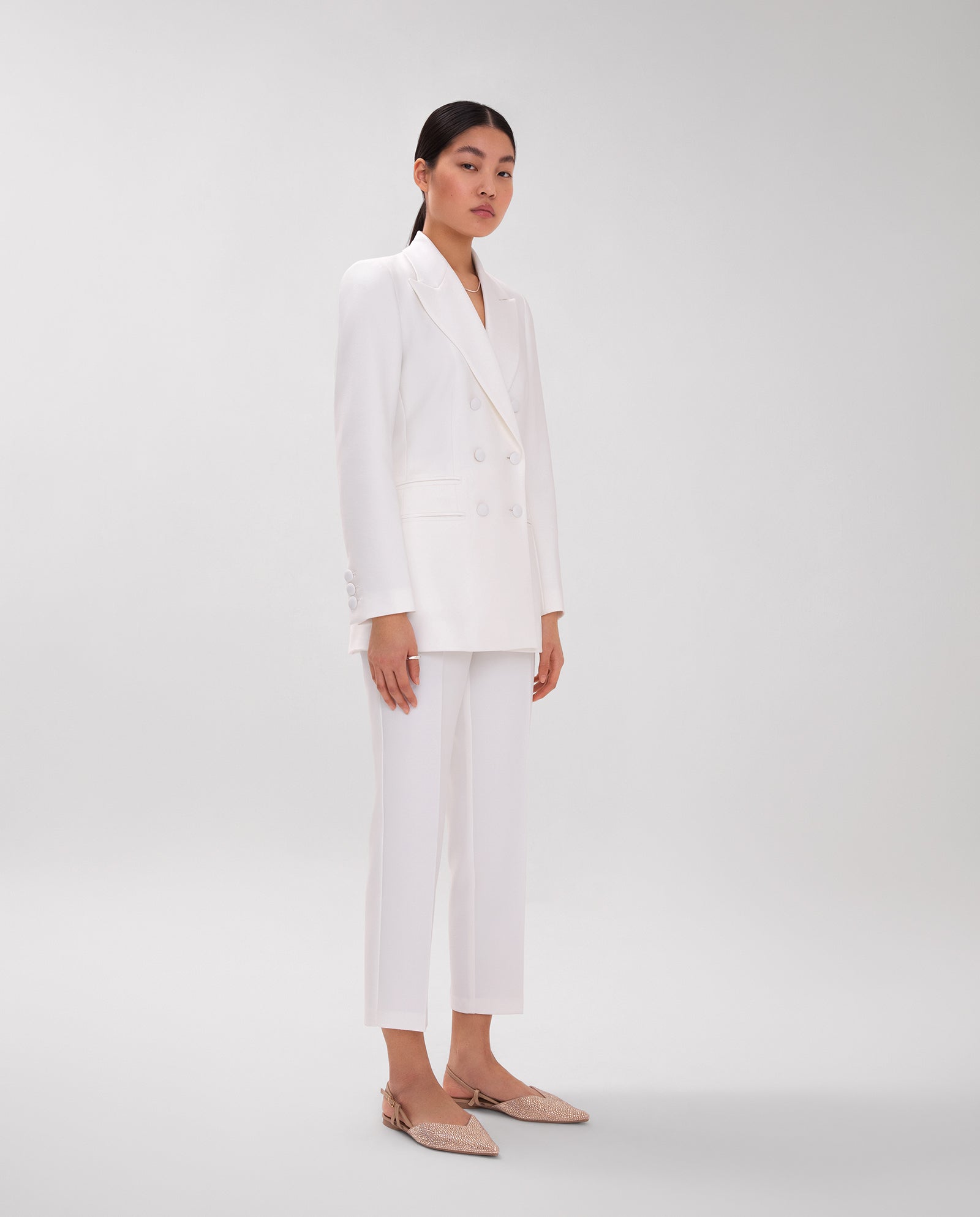 A woman wears the IVY OAK JENNIFER Blazer with a wide lapel collar, styled as a white pantsuit and paired with beige shoes, standing against a plain light-gray background.