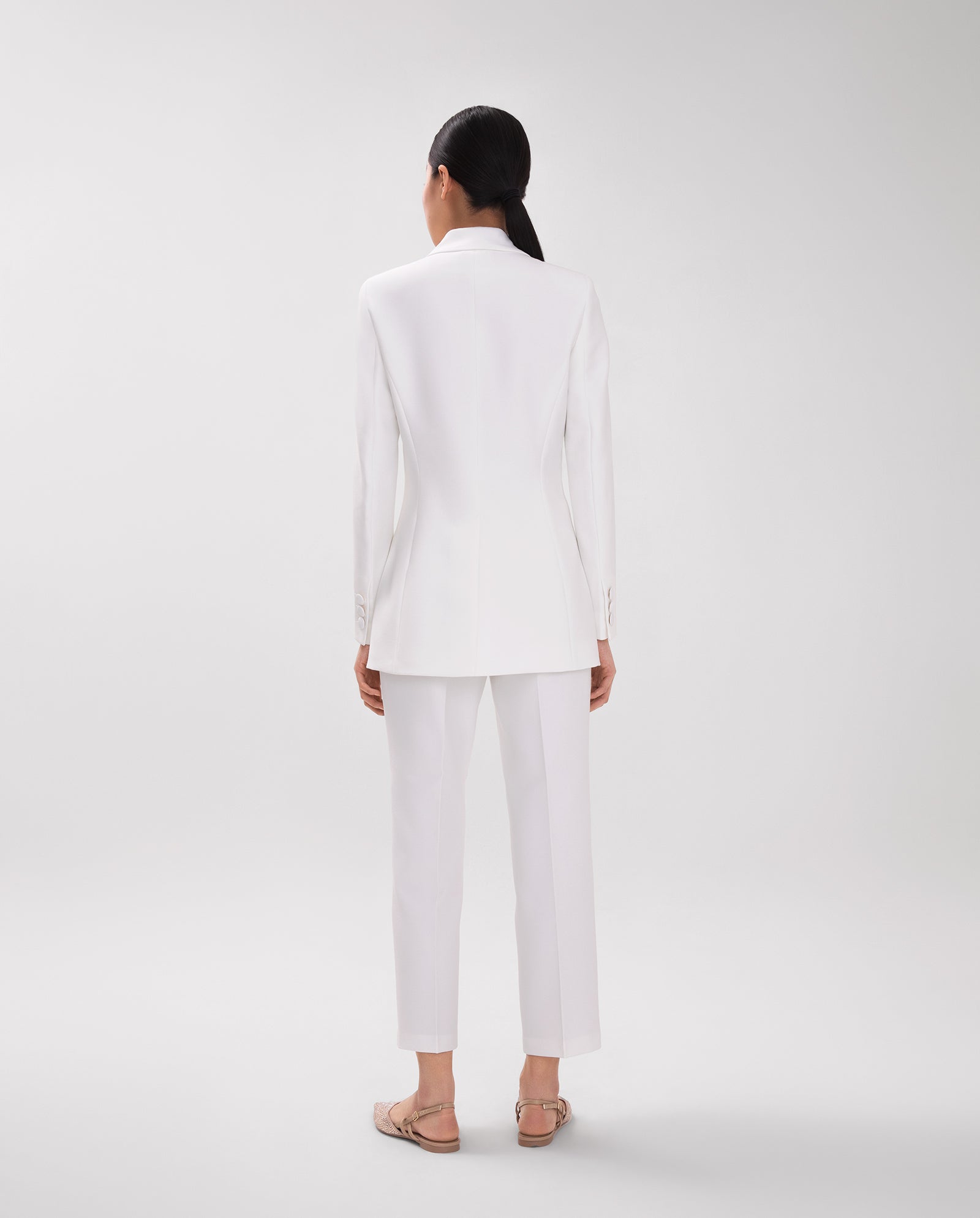 A woman stands facing away in a minimal, light studio, wearing the IVY OAK JENNIFER Blazer—a white double-breasted piece with wide lapel collar and sleeve slits—paired with sandals.