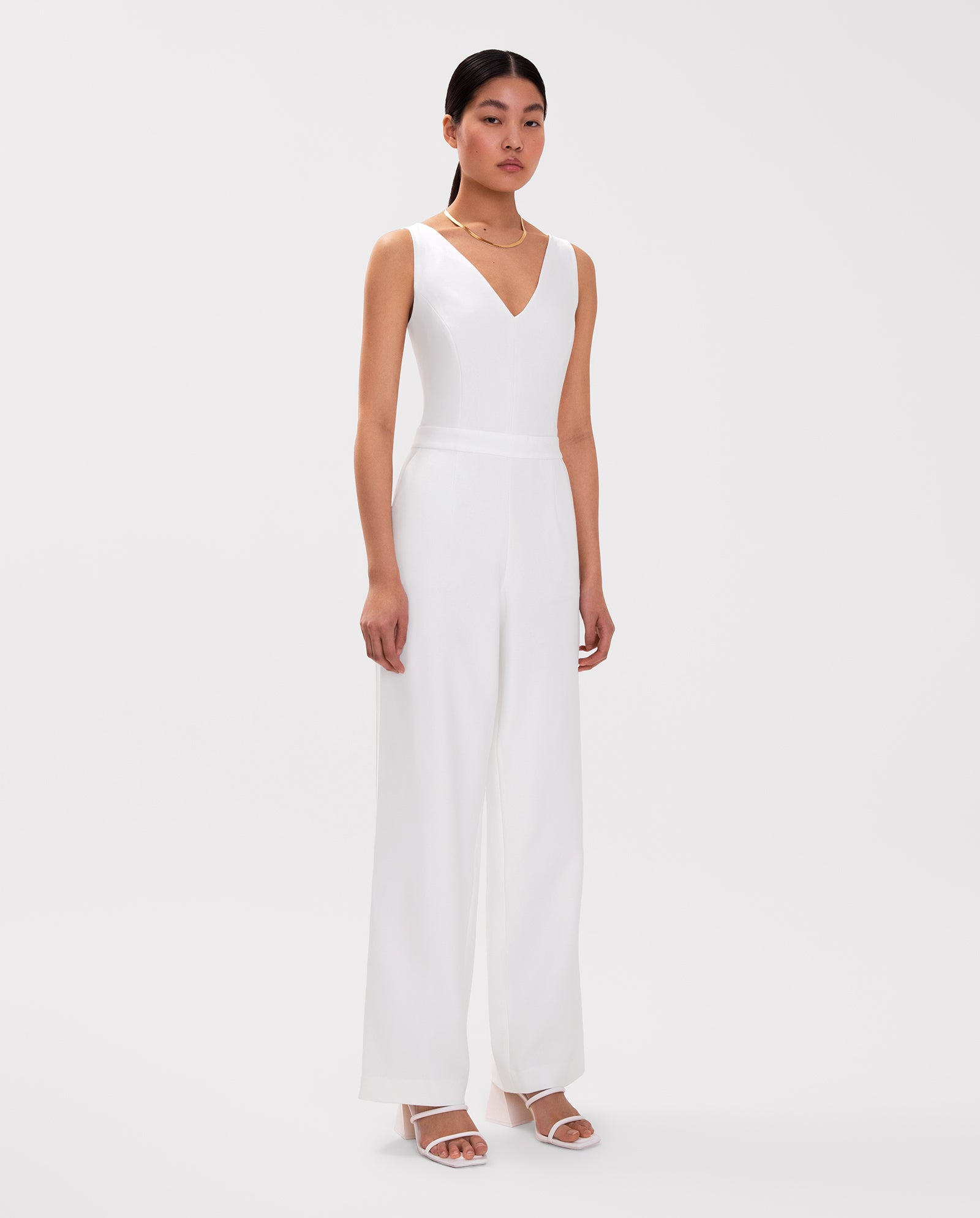 A woman stands against a plain background wearing the IVY OAK PAULINA Jumpsuit—a sleeveless white V-neck style with wide legs and a defined waistband—paired with white sandals.