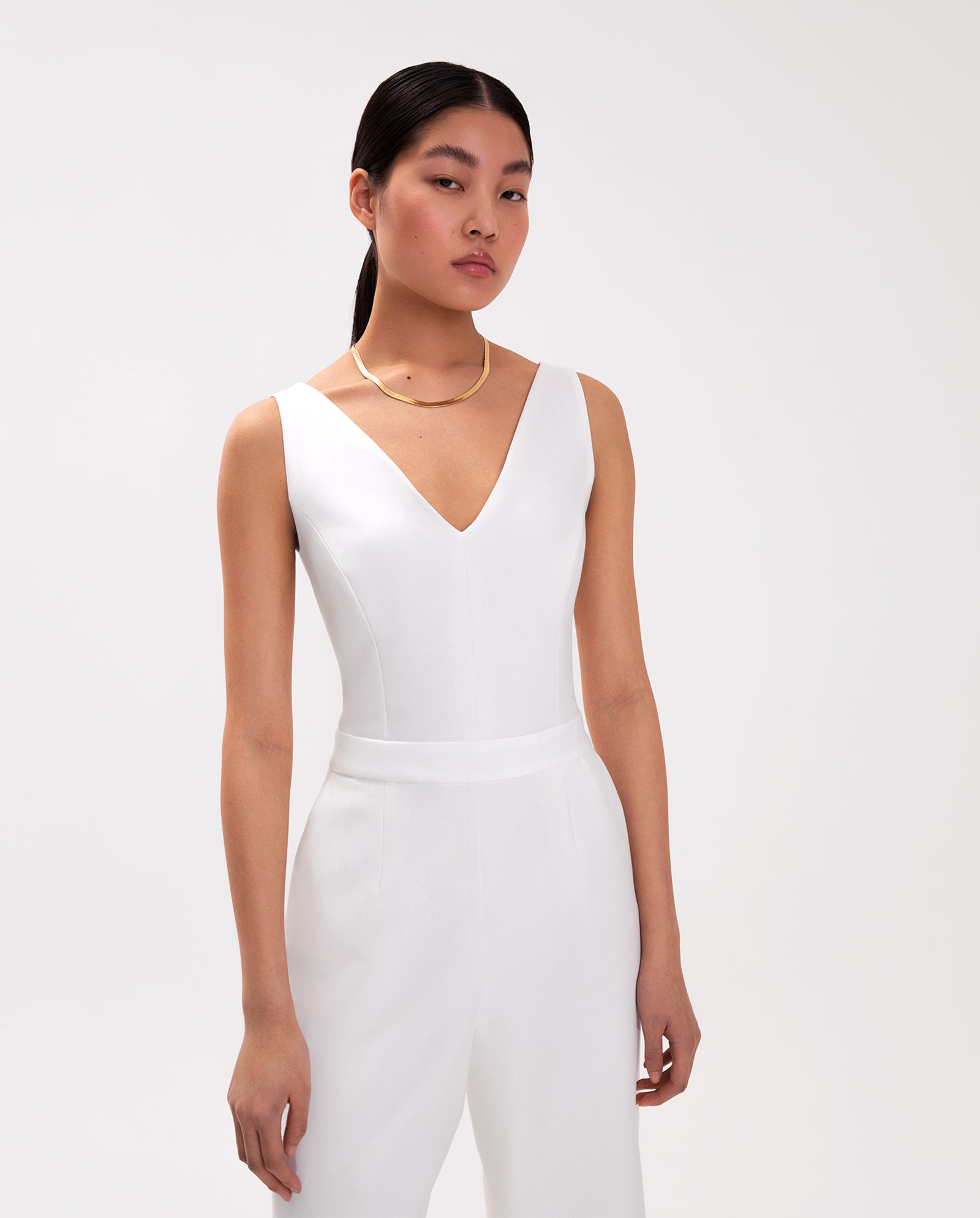 A woman wears the IVY OAK PAULINA Jumpsuit, a sleeveless white V-neck style with a defined waistband, paired with a gold necklace, standing against a white background.