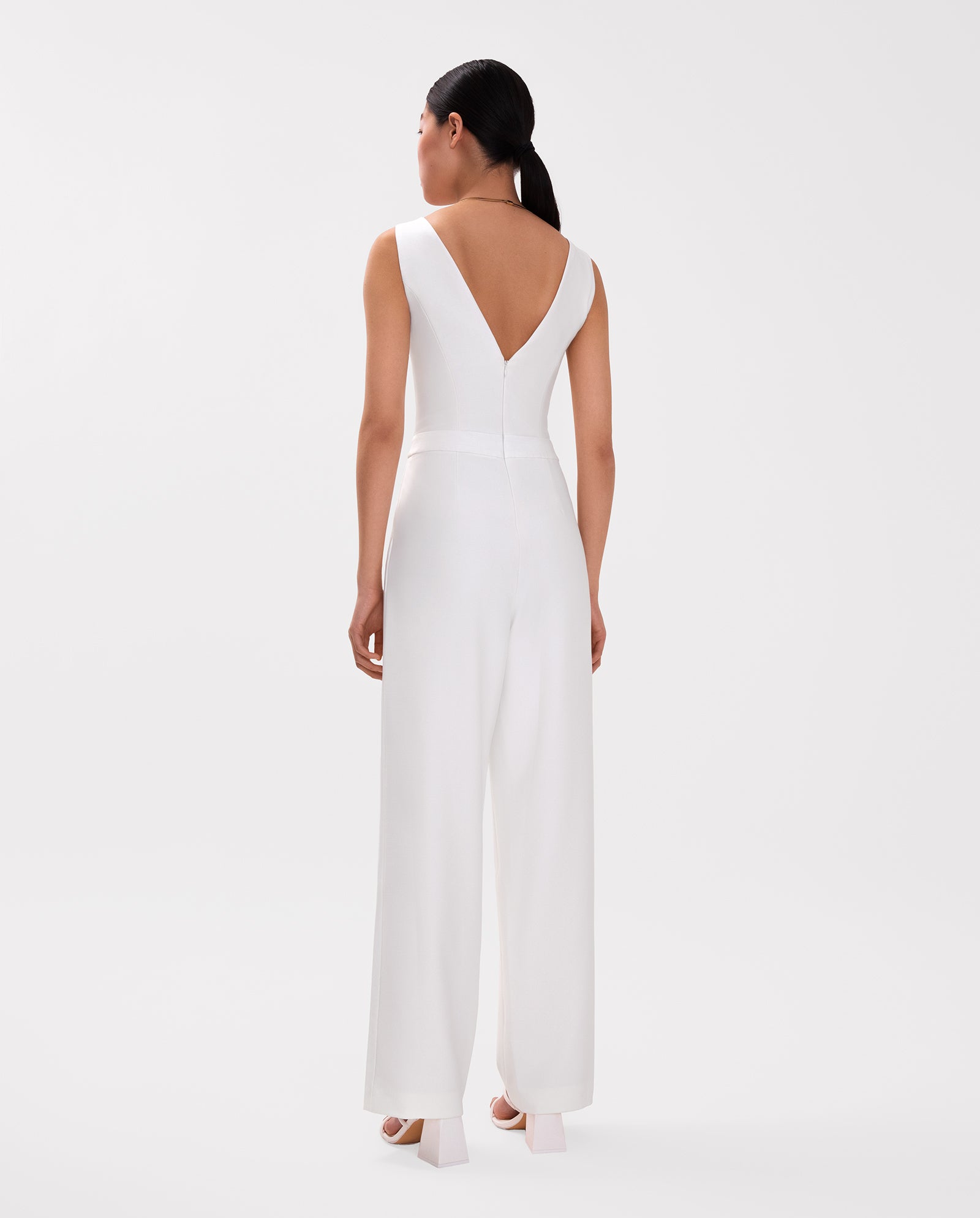Woman in the IVY OAK PAULINA Jumpsuit—a sleeveless white V-neck one-piece with a defined waistband—stands facing away against a plain background.