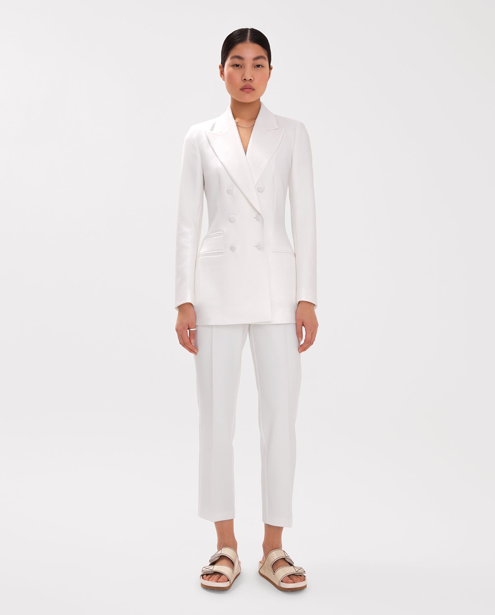 A woman wearing IVY OAKs Ankle Length PIPPA Pants and a white double-breasted suit with sandals stands against a plain white background.