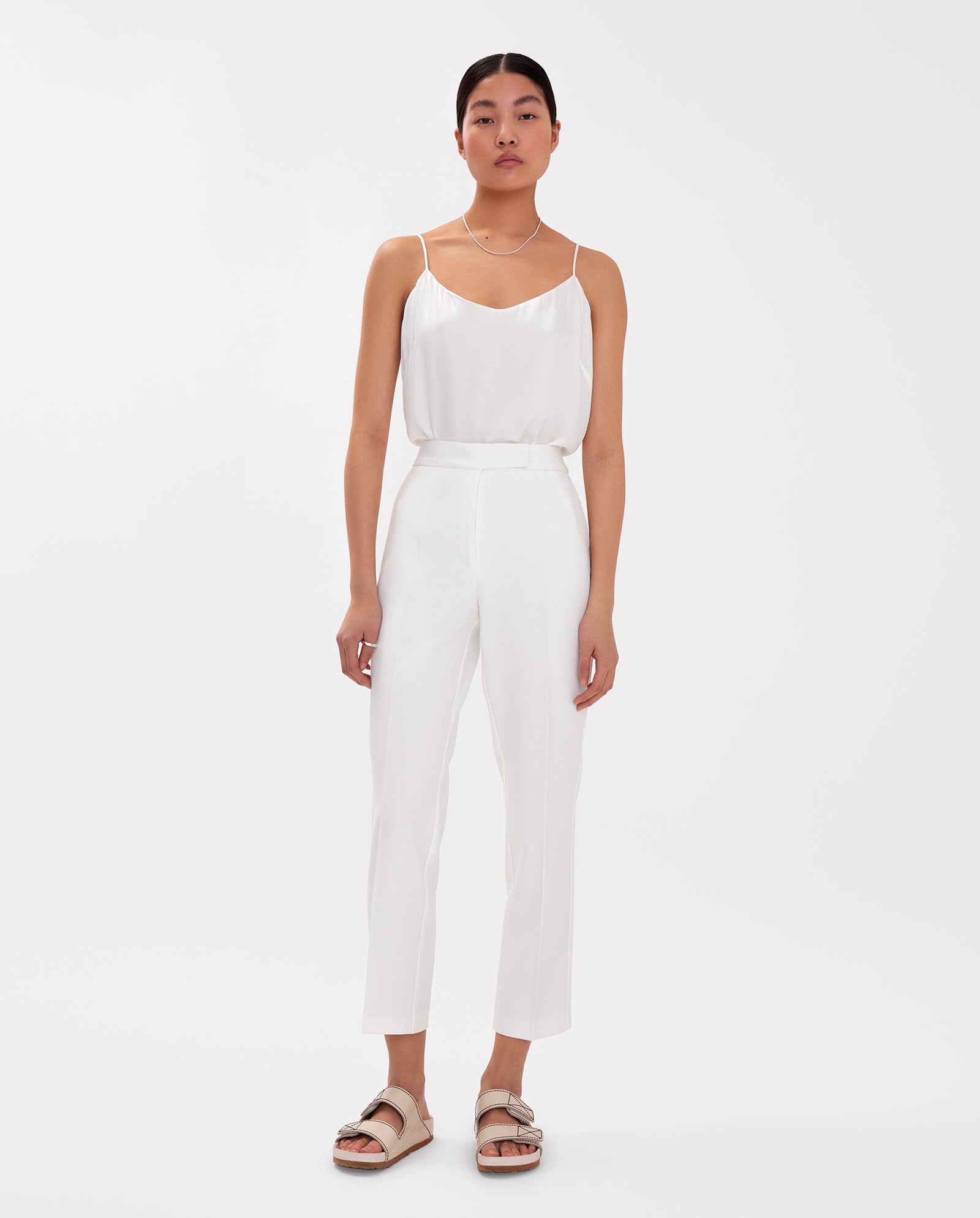 A woman stands against a white background wearing IVY OAKs Ankle Length PIPPA Pants in white, paired with a white camisole and beige sandals.