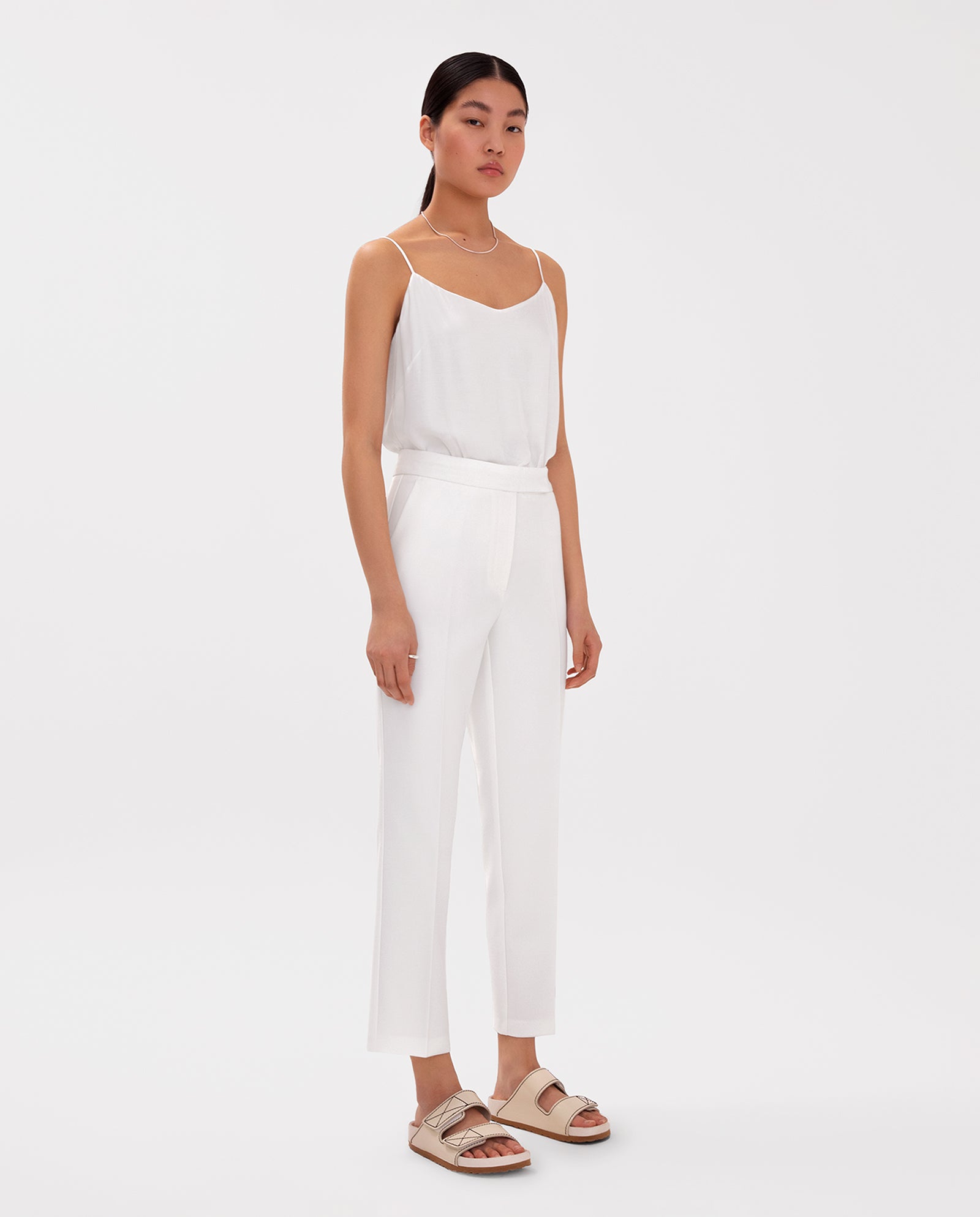A woman wearing IVY OAKs Ankle Length PIPPA Pants in white, paired with a white sleeveless top and beige sandals, stands against a white background.