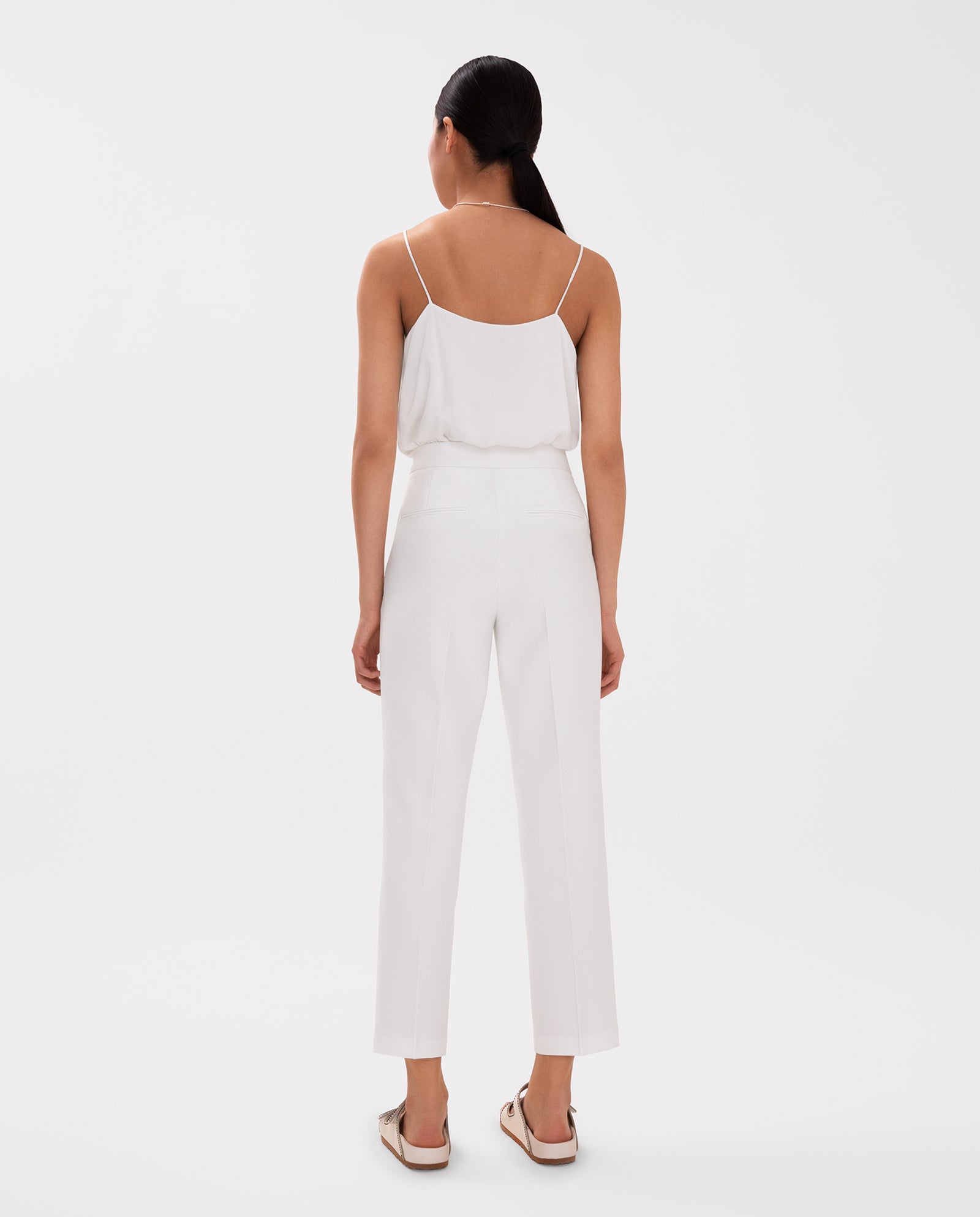 A woman faces away from the camera, wearing IVY OAK’s Ankle Length PIPPA Pants with pockets and a zipper, styled with a white sleeveless top and sandals against a white background.