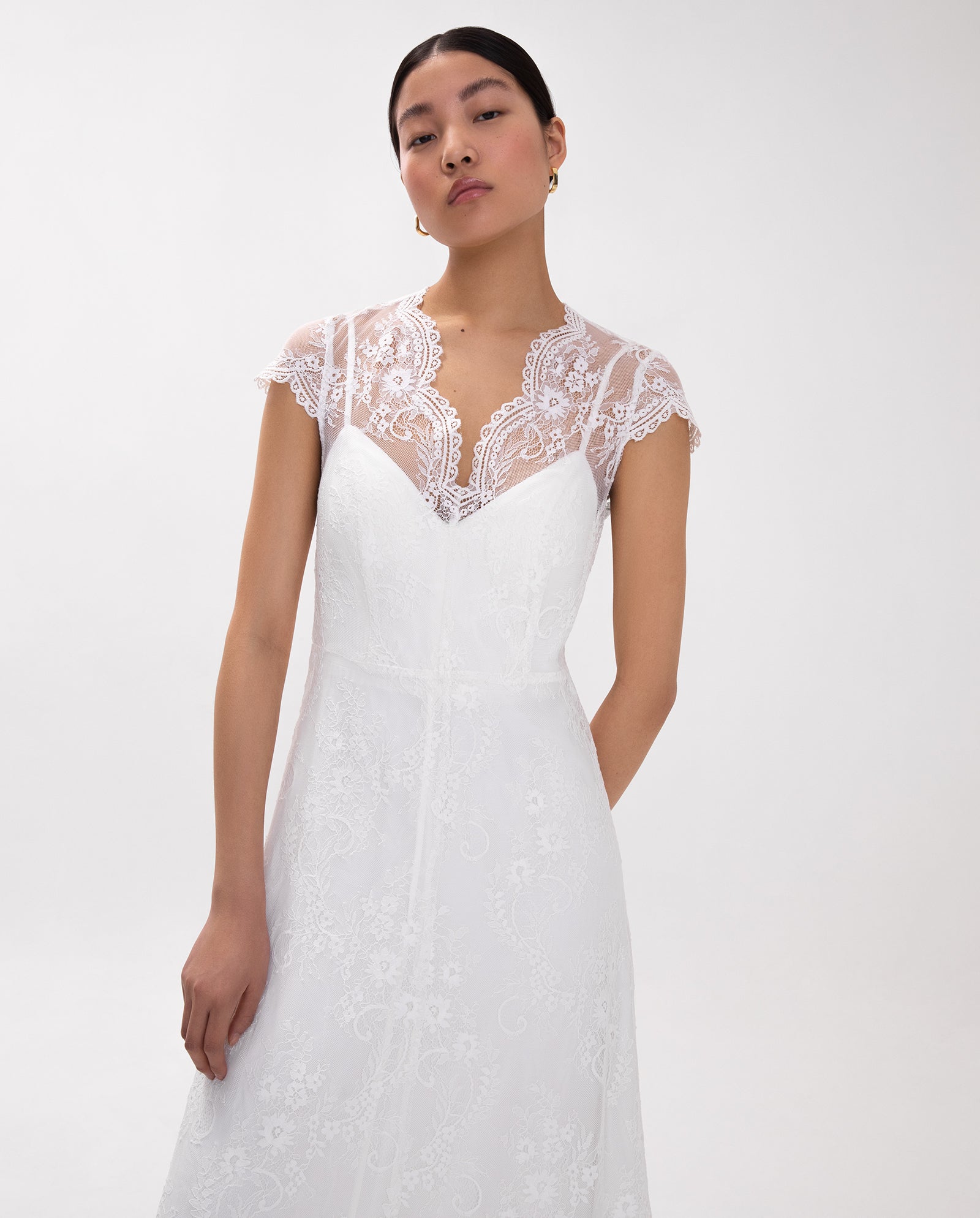Woman wearing the IVY OAK DANIELLA ROSE bridal dress, featuring white lace, an eyelash neckline, and cap sleeves, standing against a plain white background.