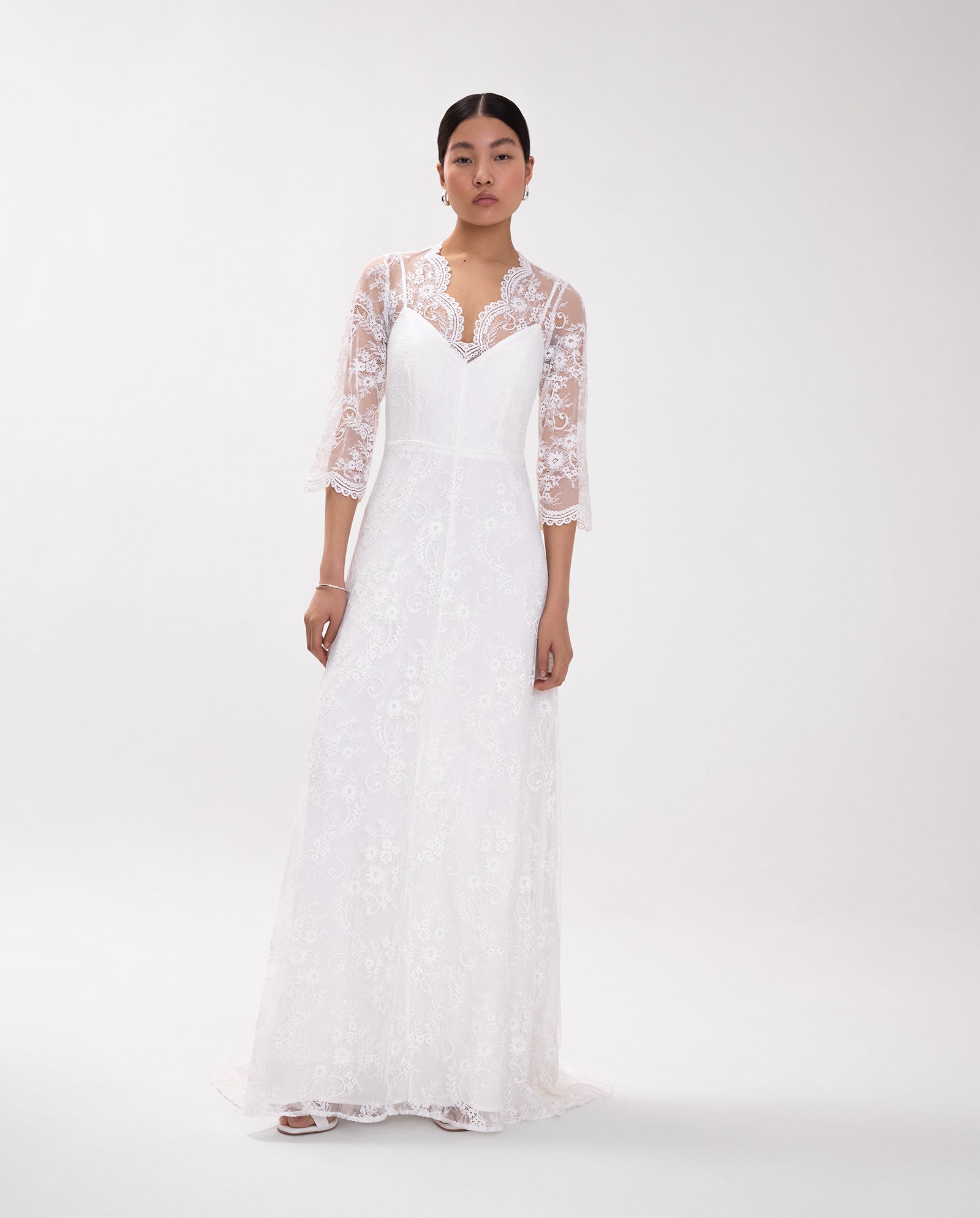 A woman models the DALIA Lace Dress by IVY OAK, featuring a long white lace design with three-quarter sleeves and a V-neckline, set against a plain background.