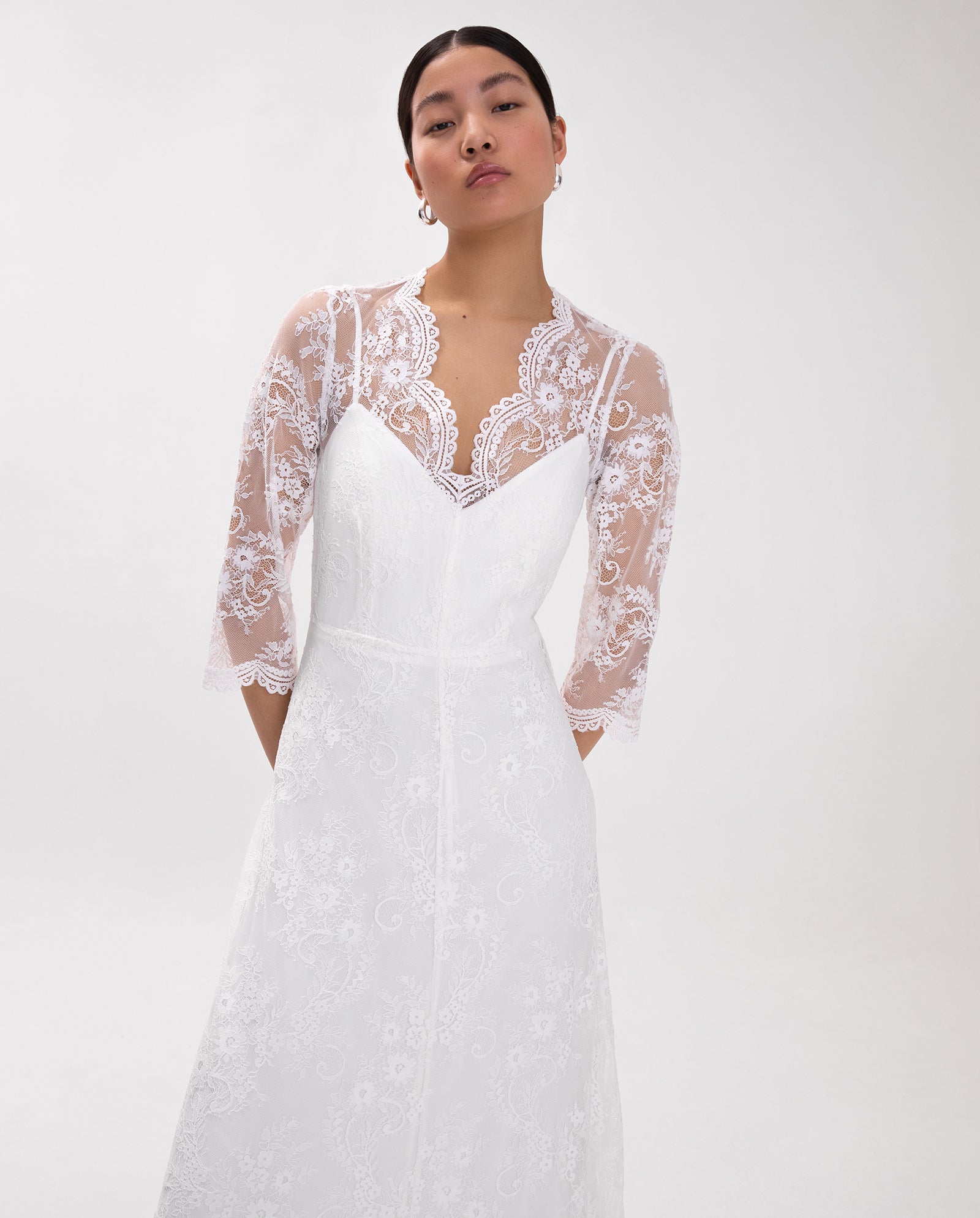 A woman in the IVY OAK DALIA Lace Dress, featuring sheer lace sleeves and a V-neckline, stands against a plain light background.
