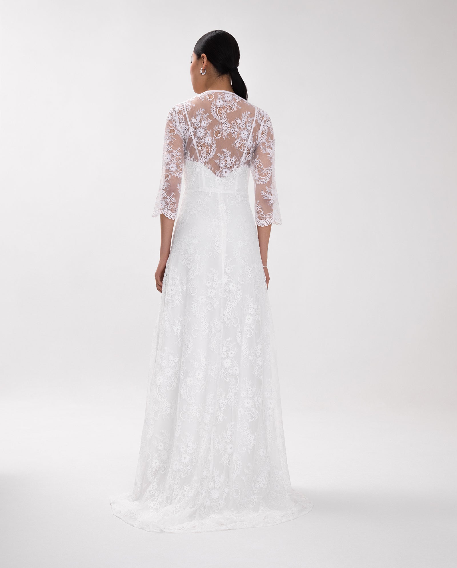 Woman in the IVY OAK DALIA Lace Dress, a long white gown with sheer sleeves and an elegant V-neckline, shown from the back against a plain background.