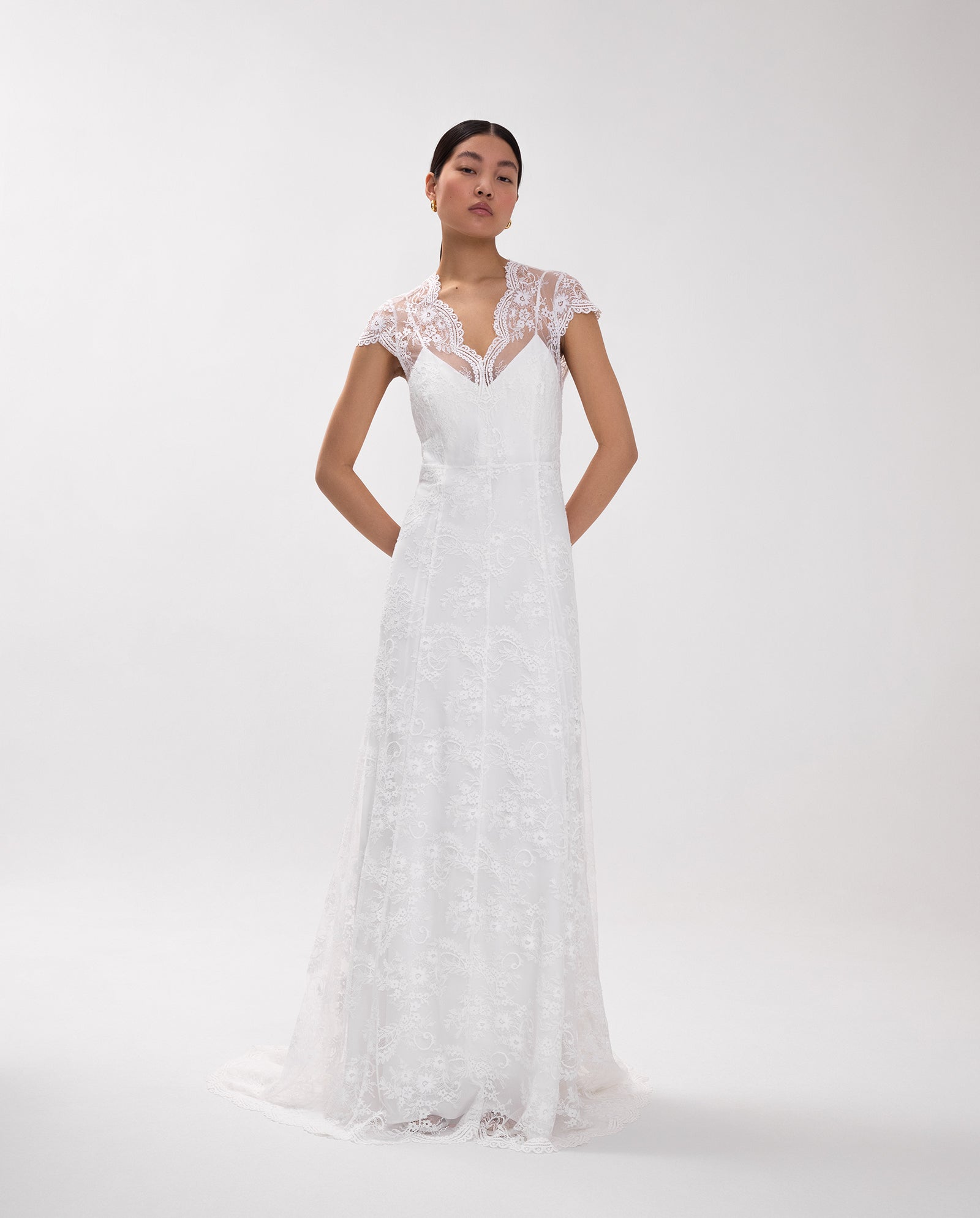 A woman wears the IVY OAK DANIELA LILY Dress, a white maxi bridal gown featuring delicate lace cap sleeves and a high neckline, standing against a plain light background.