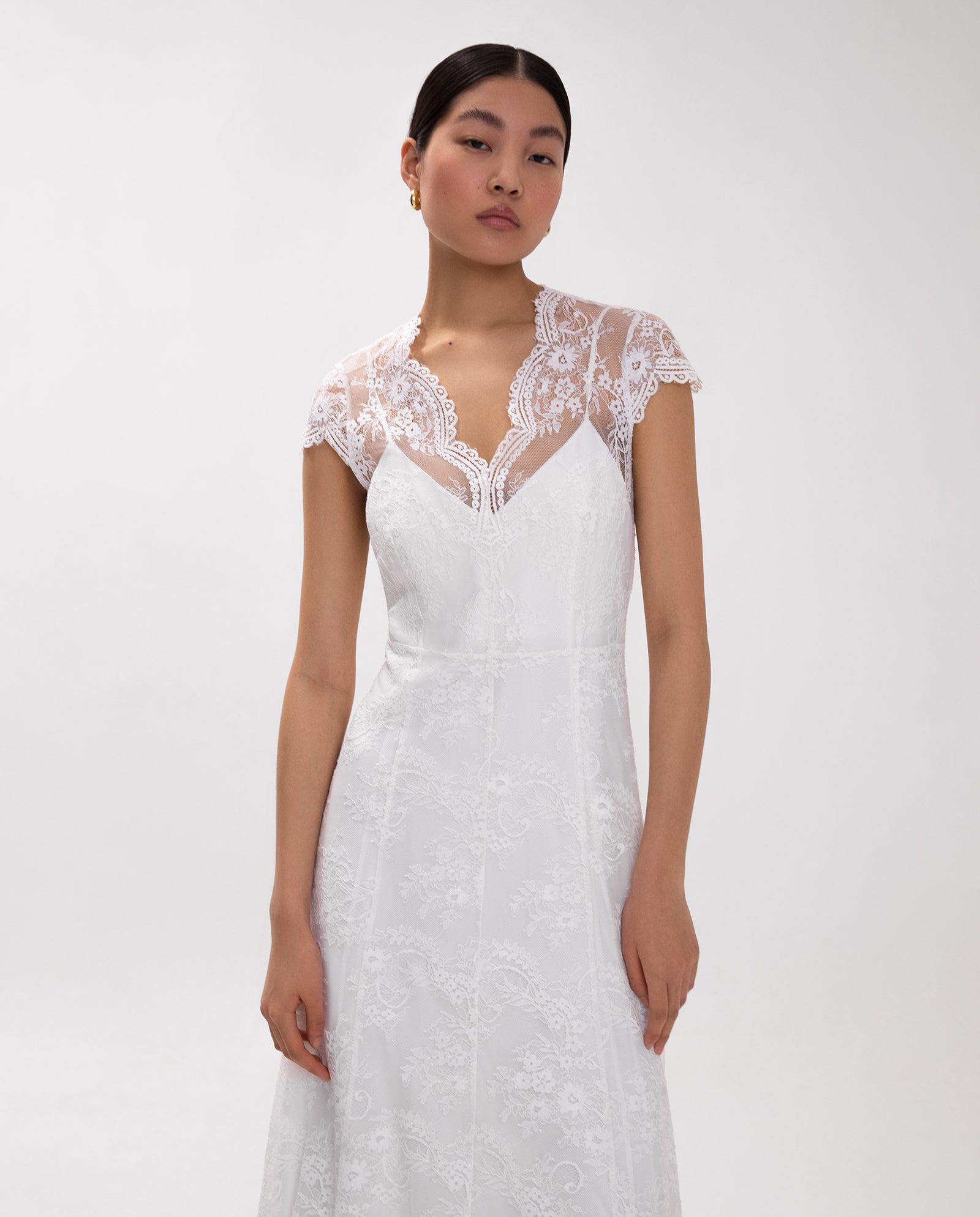 A woman wearing the IVY OAK DANIELA LILY white lace V-neck maxi bridal dress with cap sleeves stands against a plain white background.