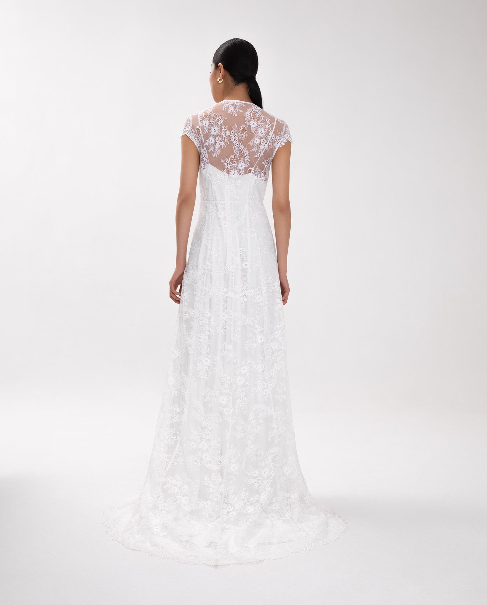 Woman wearing the DANIELA LILY Dress by IVY OAK, a white lace V-neck wedding gown with a sheer floral back and adjustable straps, stands facing away against a plain background.