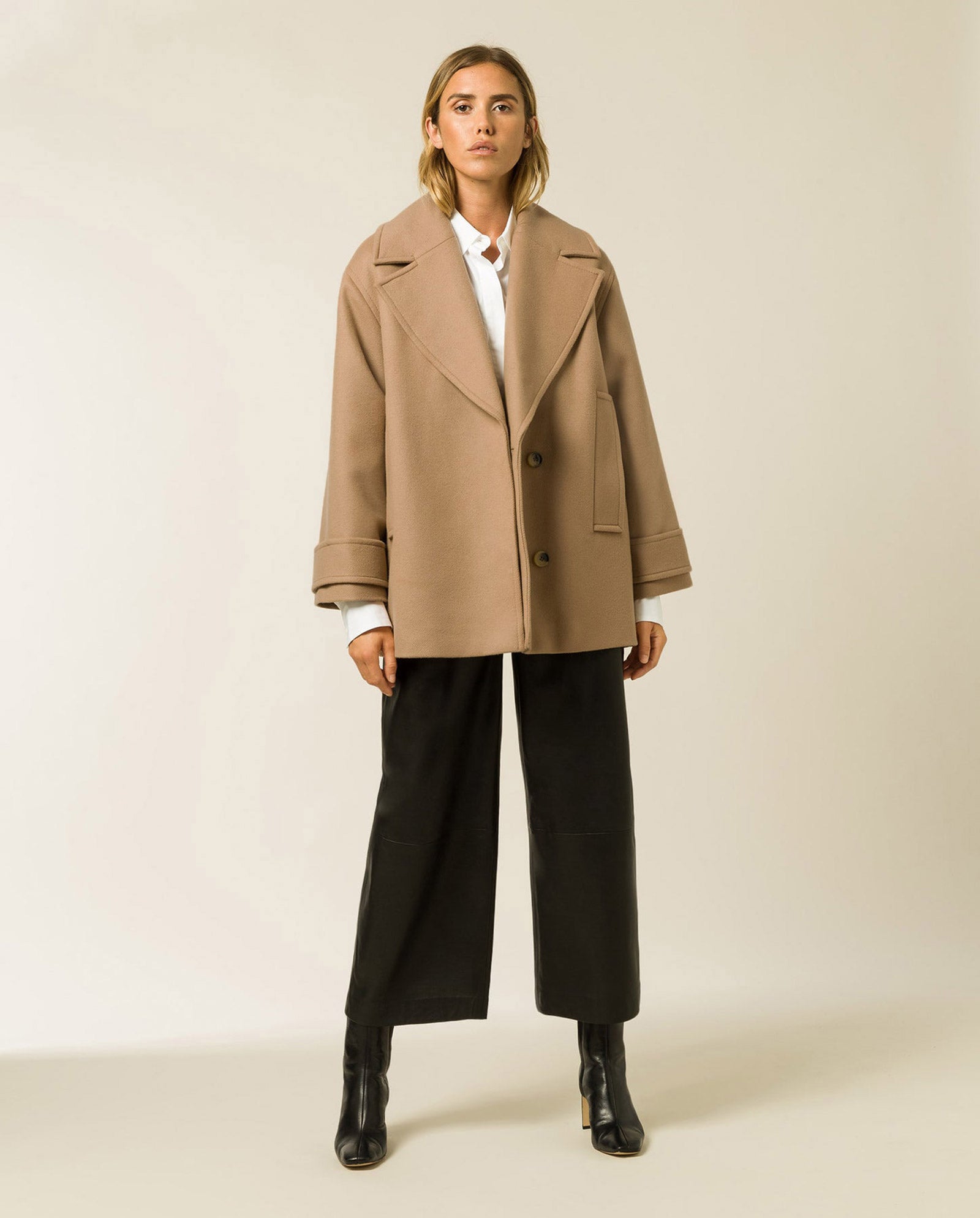 A woman wears the CARLY Wool Coat by IVY OAK—an oversized-fit winter pea coat in Italian wool—paired with a white shirt, black wide-leg pants, and black ankle boots.