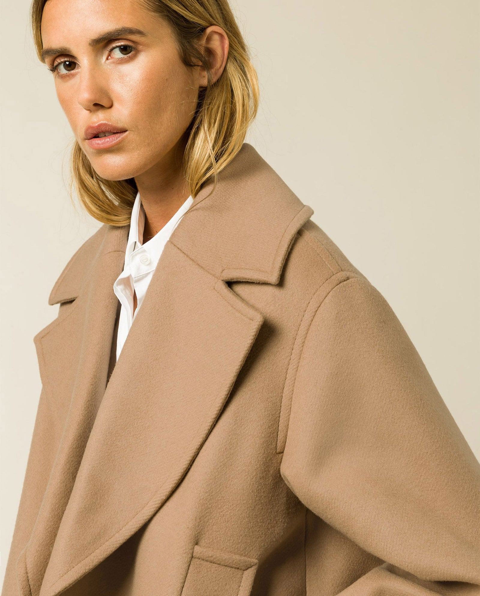 A woman models the IVY OAK CARLY Wool Coat in tan, featuring an oversized fit, over a white shirt while looking at the camera against a neutral background.