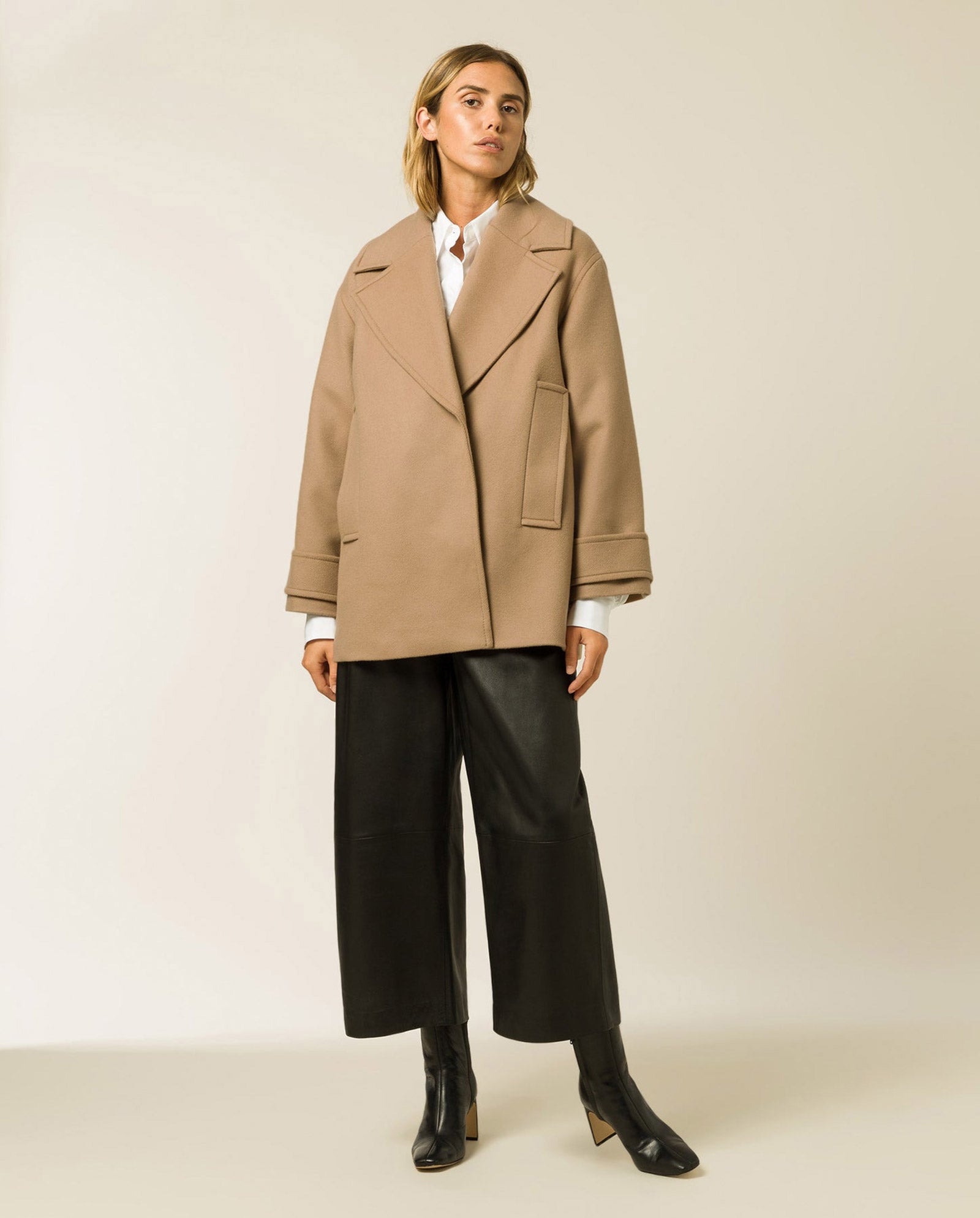 A woman wears the oversized CARLY Wool Coat in tan by IVY OAK, styled with black wide-leg pants and black boots, standing against a plain background.