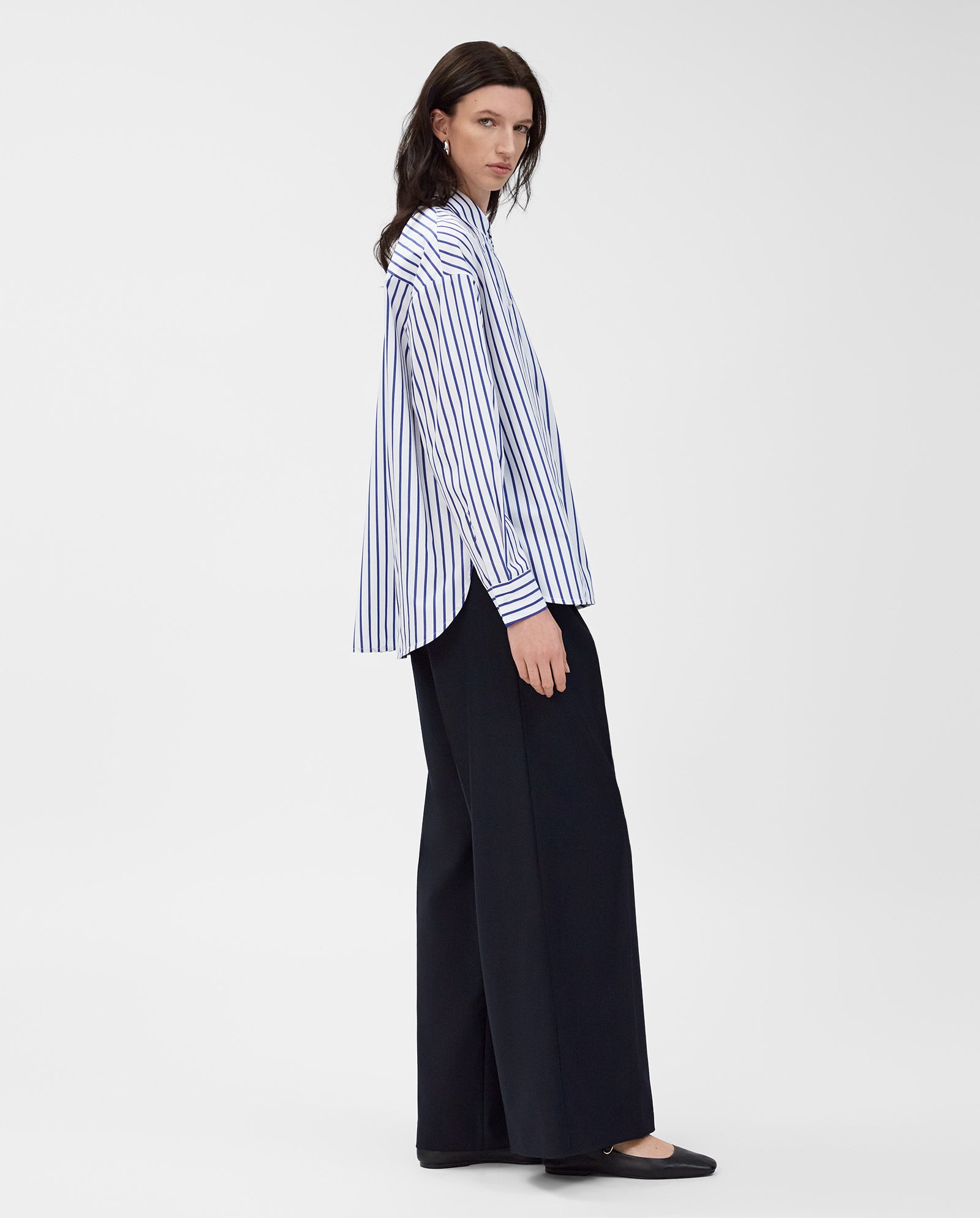 A woman stands against a white background wearing the IVY OAK BETHANY LILLY blue and white striped cotton blouse with a boxy cut, paired with black wide-leg pants.