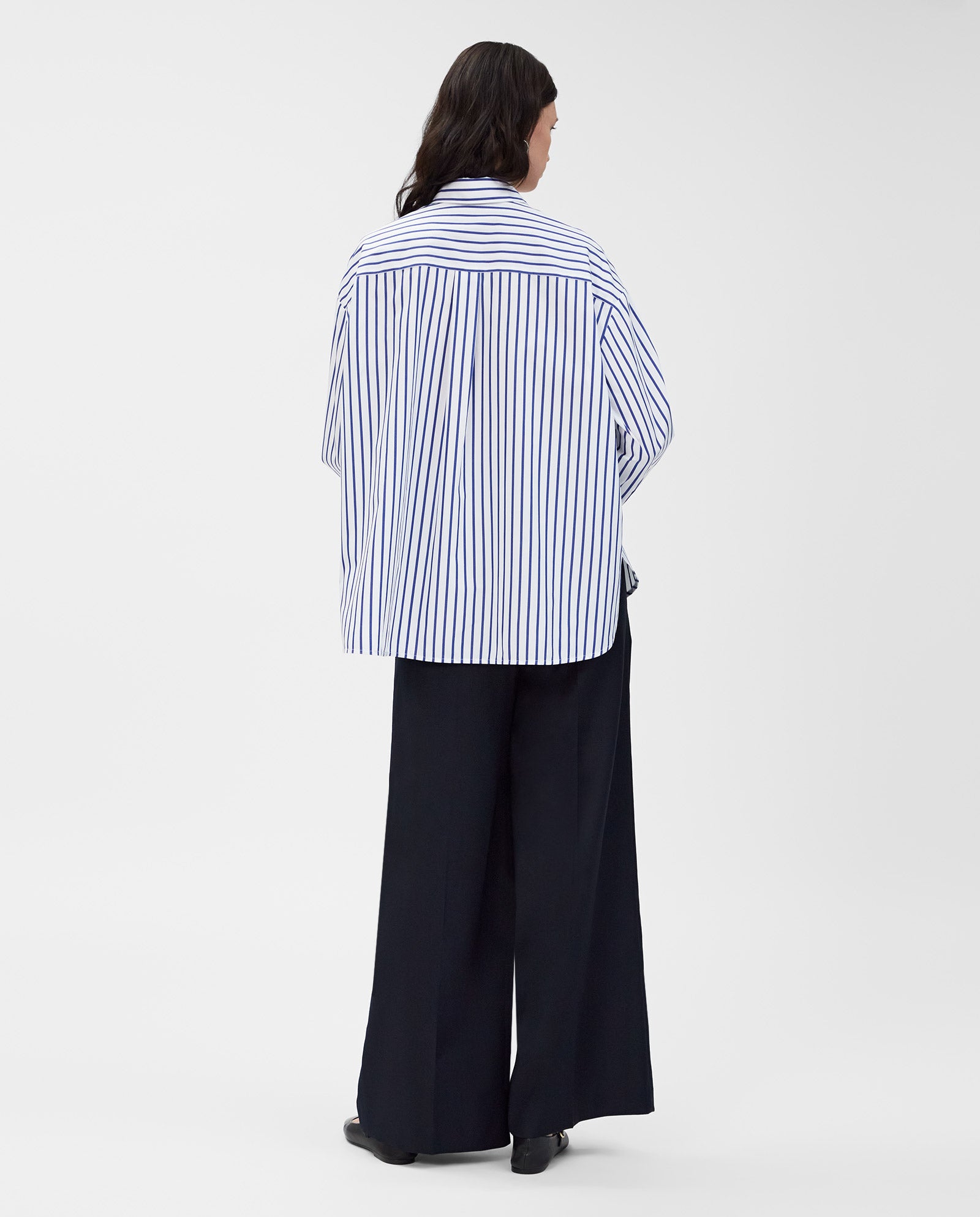 A person faces away from the camera, wearing the IVY OAK BETHANY LILLY Blouse—an oversized, striped cotton top with a boxy cut—paired with wide-leg black pants.