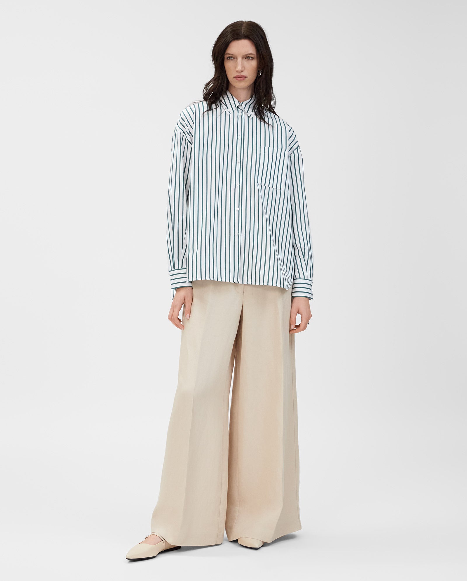 A woman stands against a white background wearing the IVY OAK BETHANY LILLY blouse, featuring a boxy cut and stripes, paired with wide-leg beige pants.